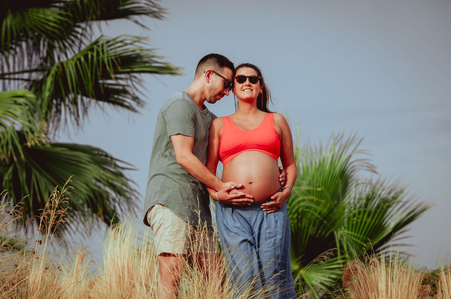 Expectant couple sharing tender moment outdoors, man leaning toward pregnant woman in red crop top near palm trees.