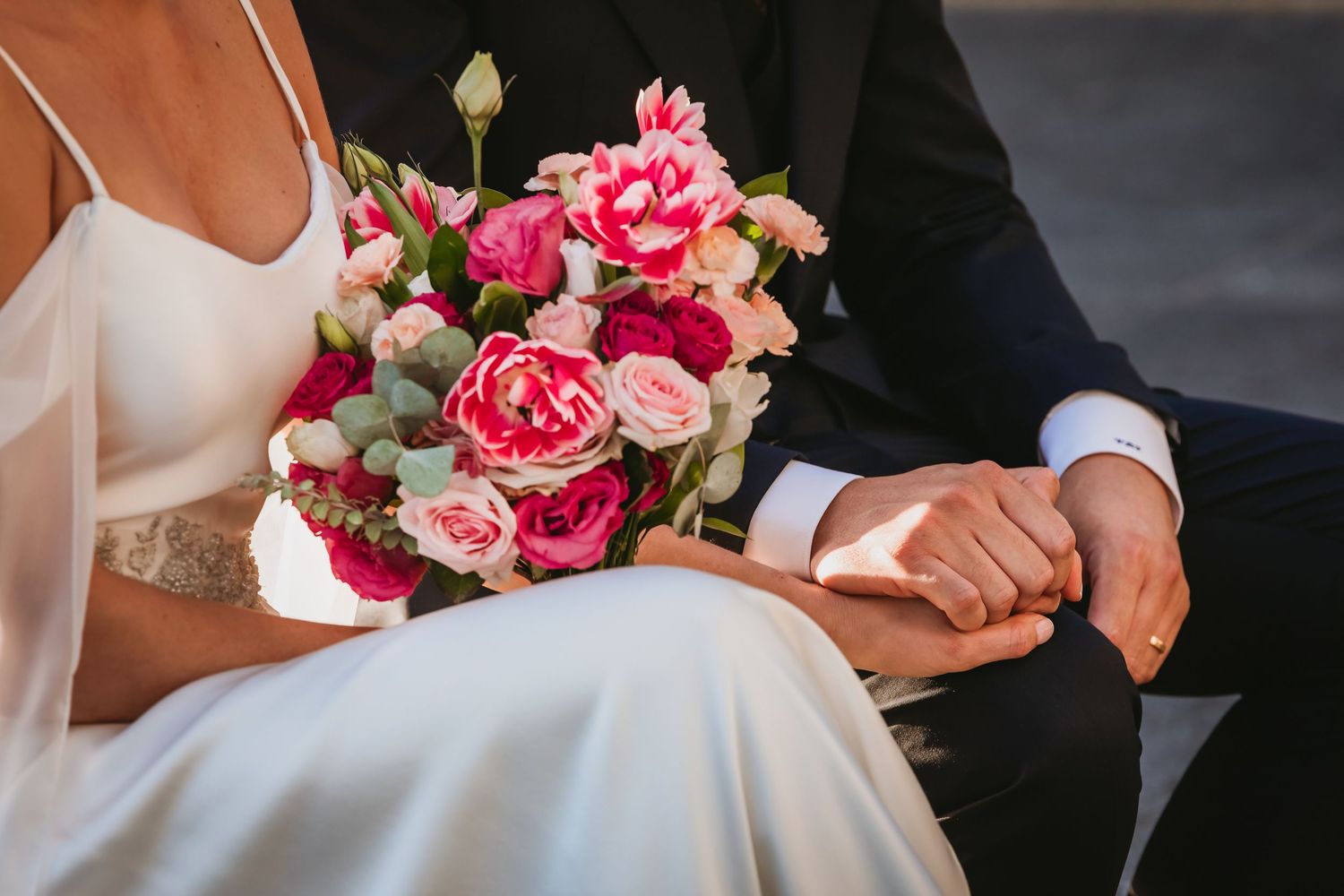 Close-up of bride and groom holding hands beside a vibrant pink and red floral bouquet.