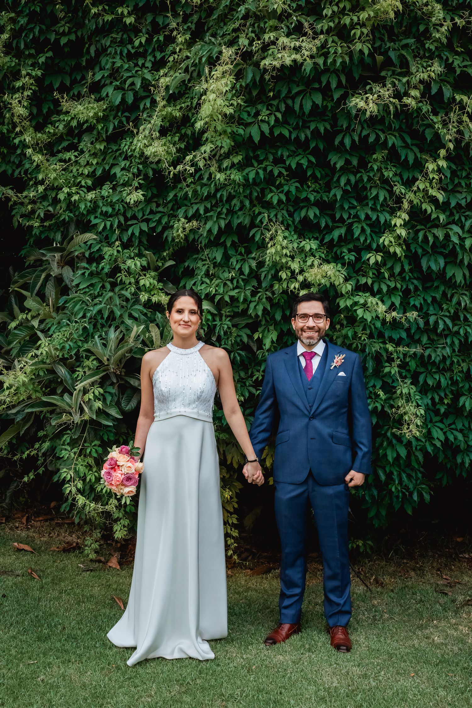 Bride in halter gown and groom in navy suit hold hands before ivy-covered wall.