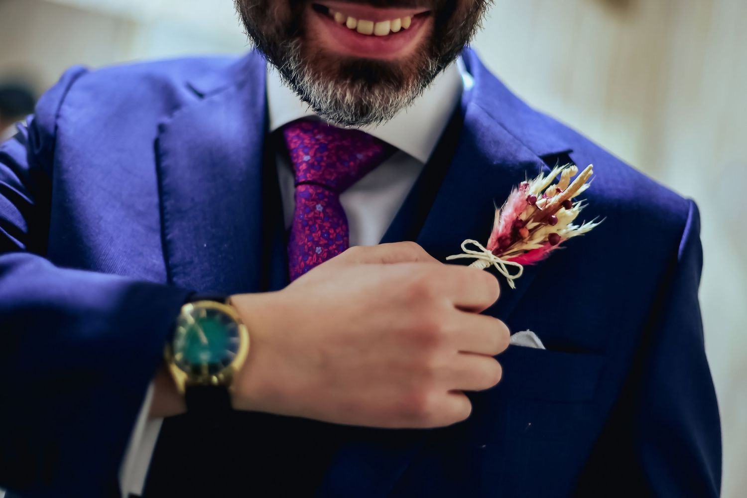 Smiling groom in blue suit with purple tie adjusting floral boutonniere while wearing gold watch.