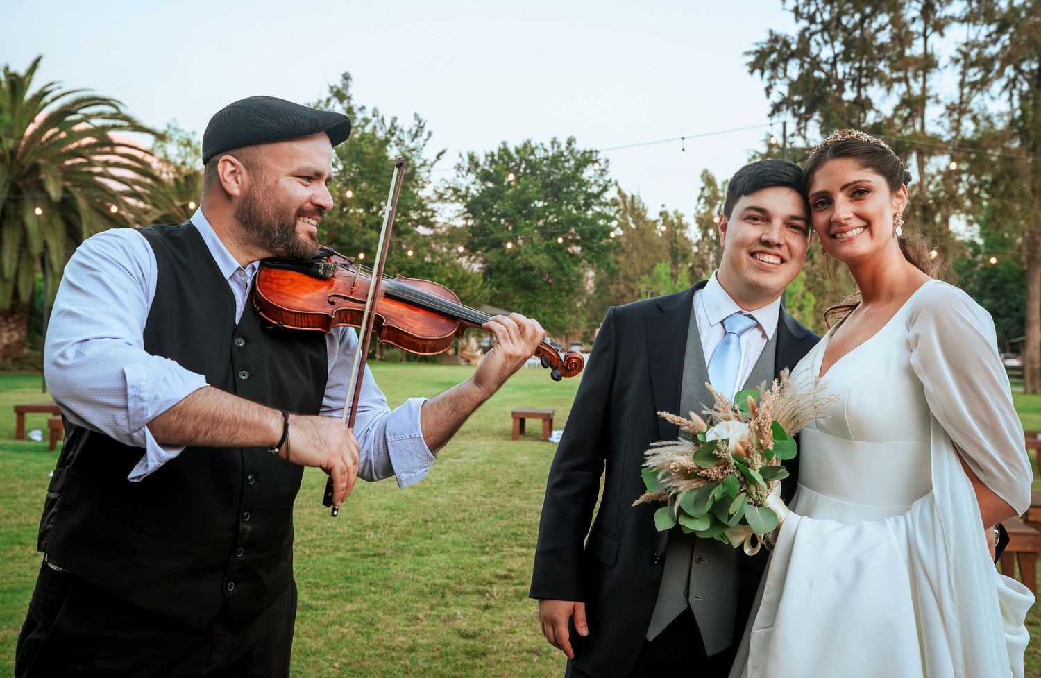 Smiling bride and groom enjoy live violin serenade from musician during outdoor wedding celebration.