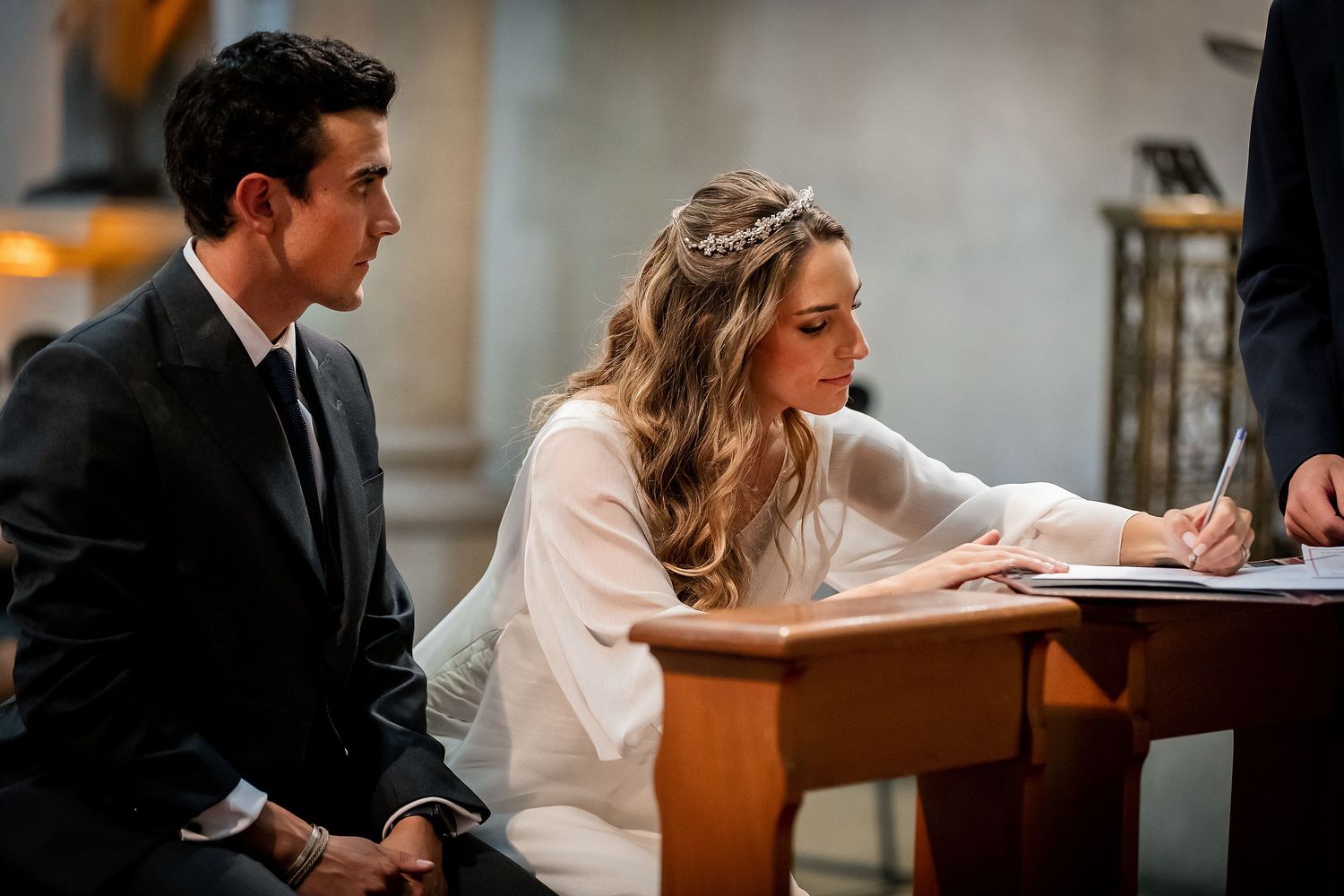 Bride signs wedding registry document while groom watches during church ceremony.