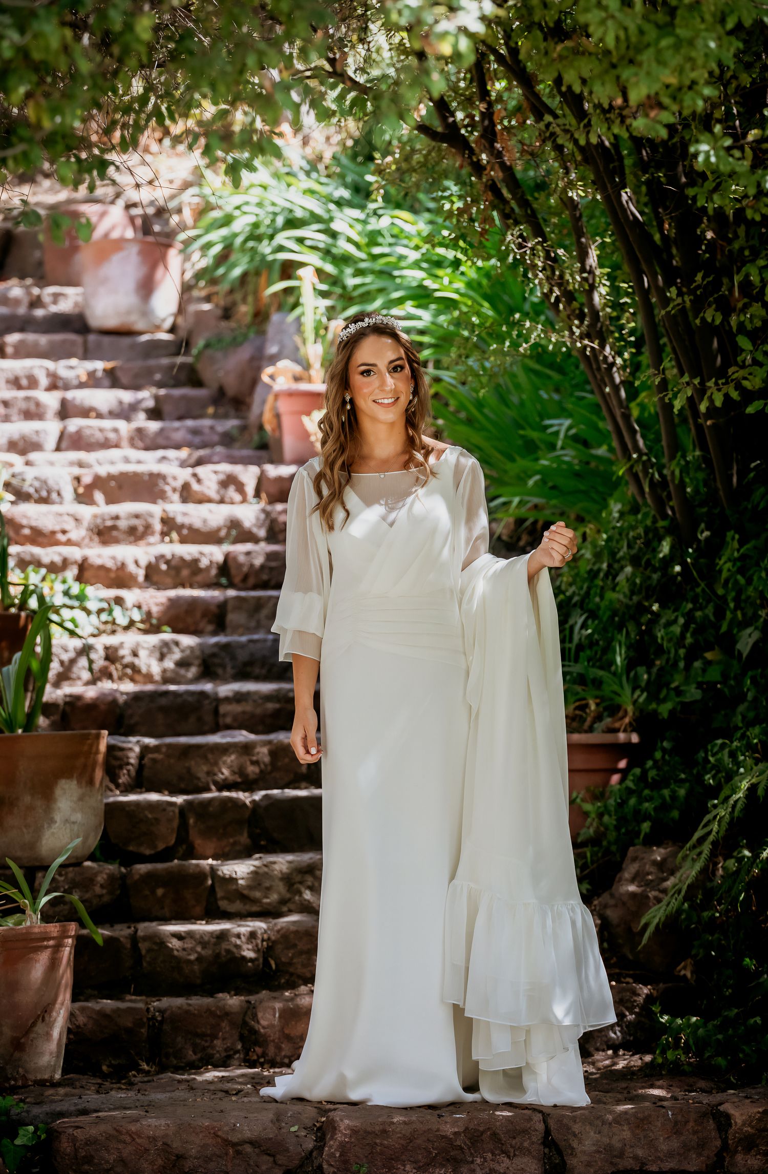 Elegant bride in a flowing white gown with sheer cape stands on stone steps in a lush garden.