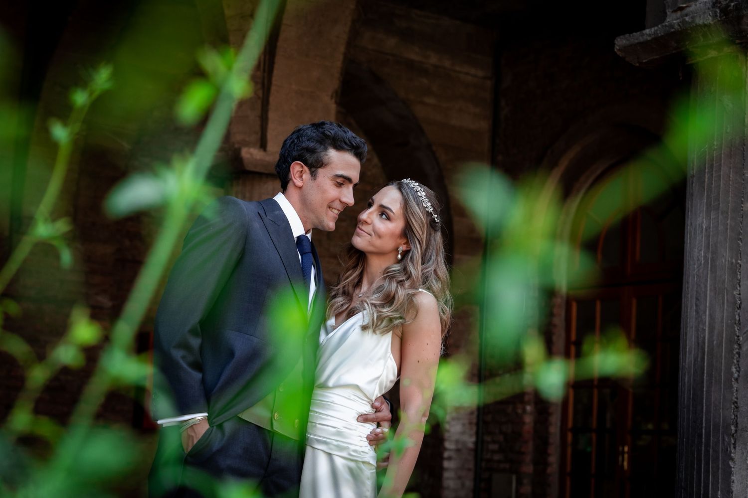 Bride and groom sharing a tender moment framed by green foliage near a rustic wooden doorway.