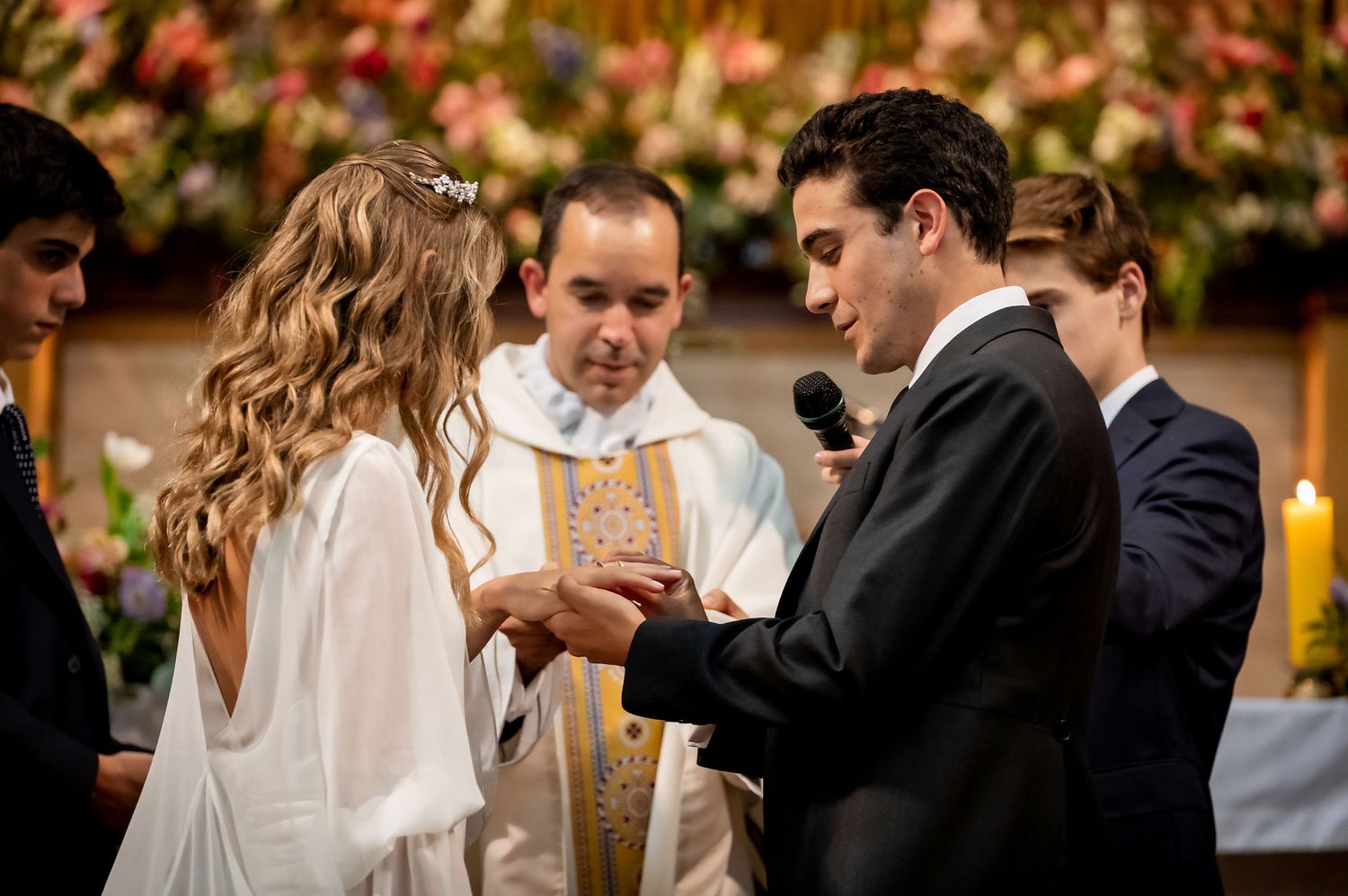 Groom placing wedding ring on brides finger during church ceremony with priest officiating.