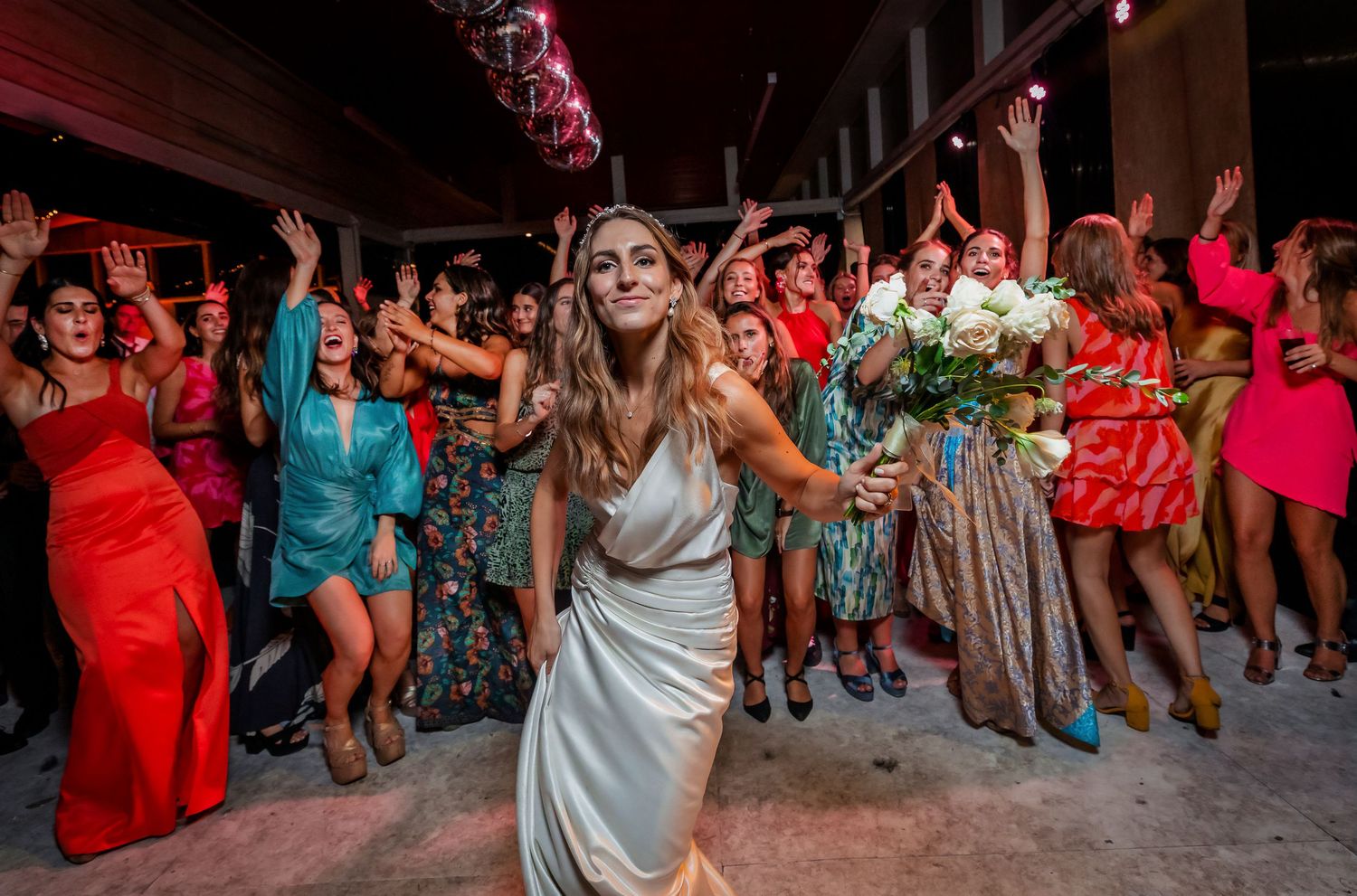 Bride in white dress prepares to toss bouquet to excited guests in colorful dresses at wedding reception.
