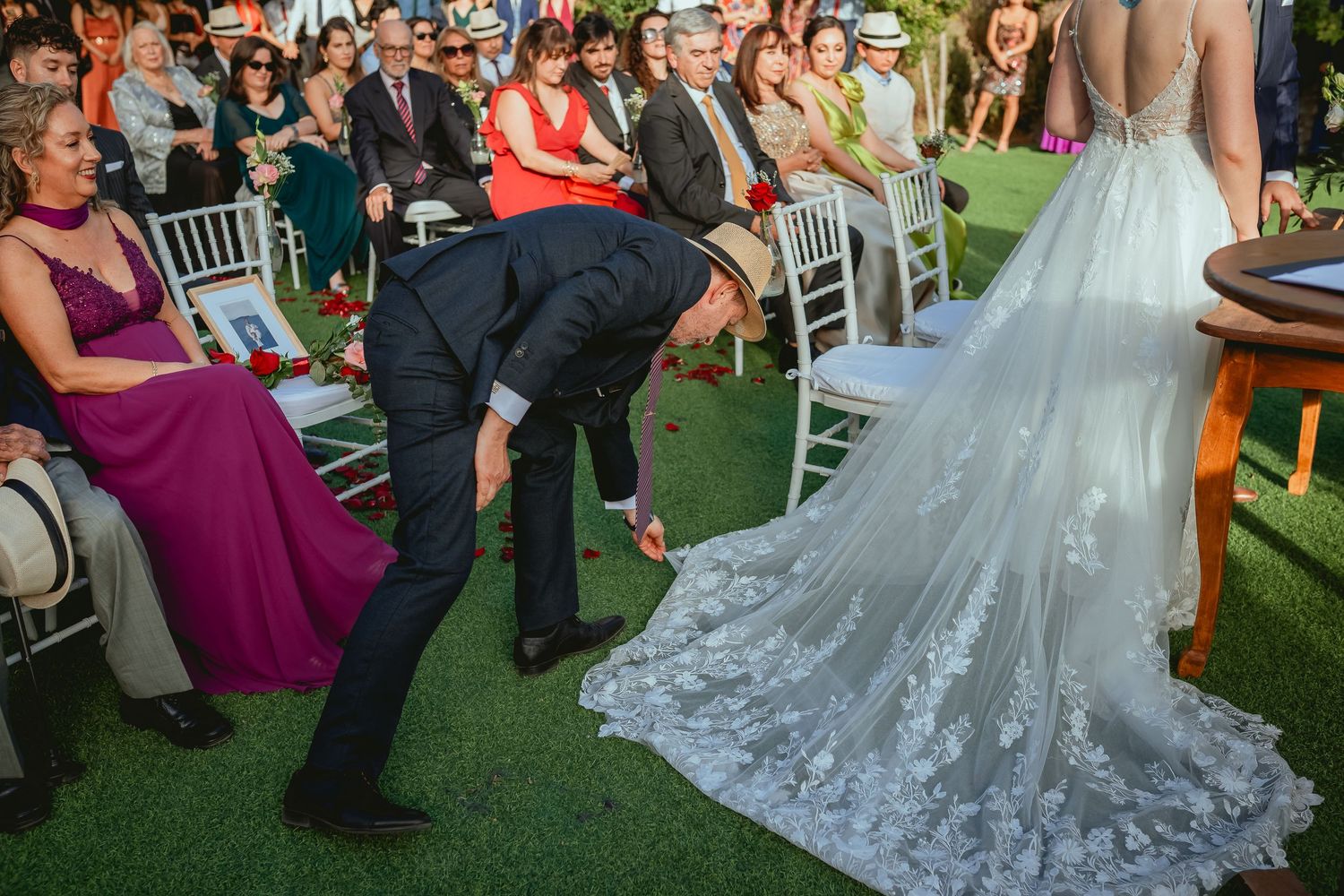 Guest adjusts bride's lace wedding dress train during outdoor garden wedding ceremony.