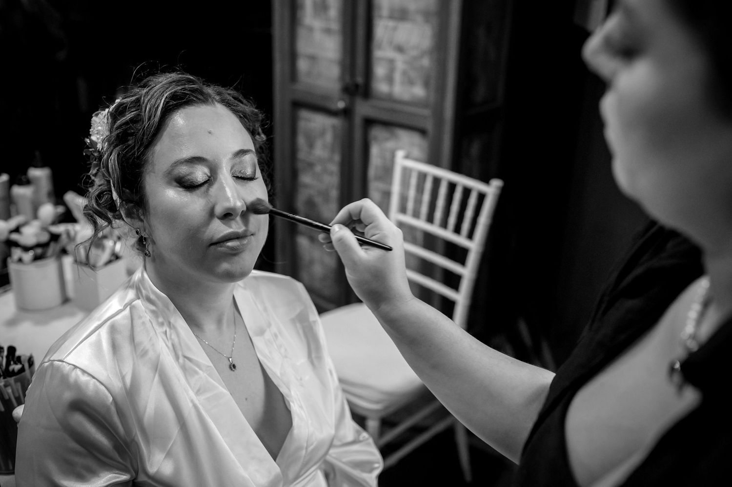 Makeup artist applying foundation to bride's face with brush during bridal preparation in black and white.