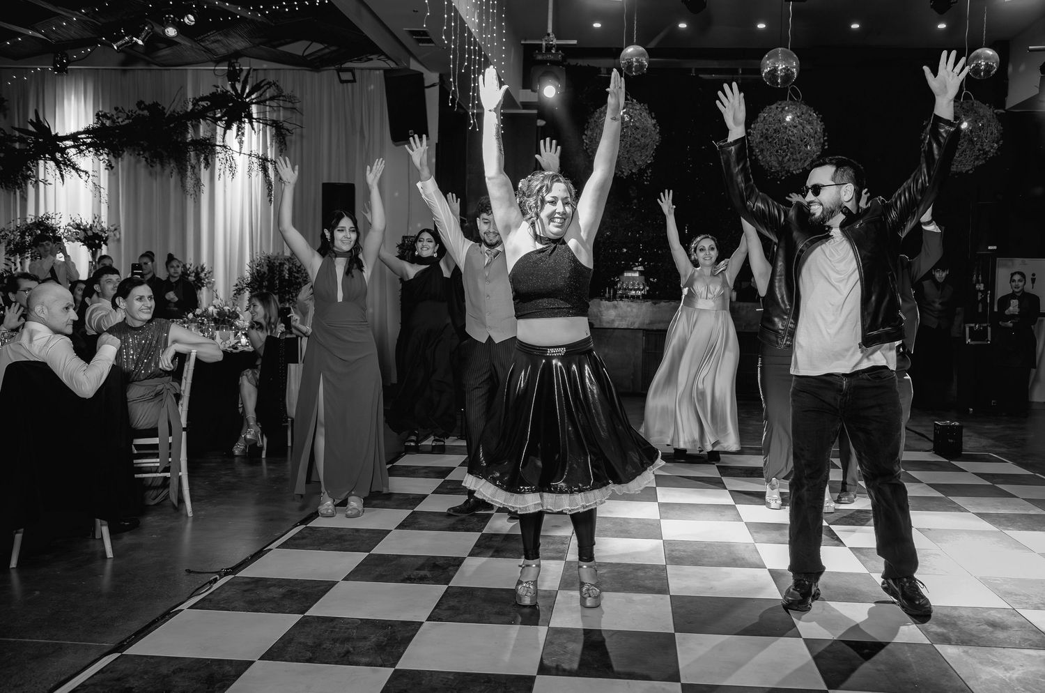 Dancers celebrate with raised arms on checkered dance floor at festive wedding reception event.