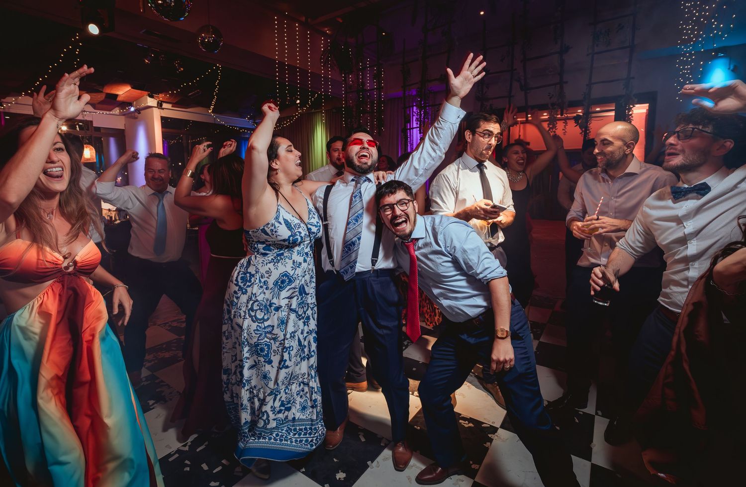 Wedding guests dance and celebrate with groom in suspenders at indoor reception.