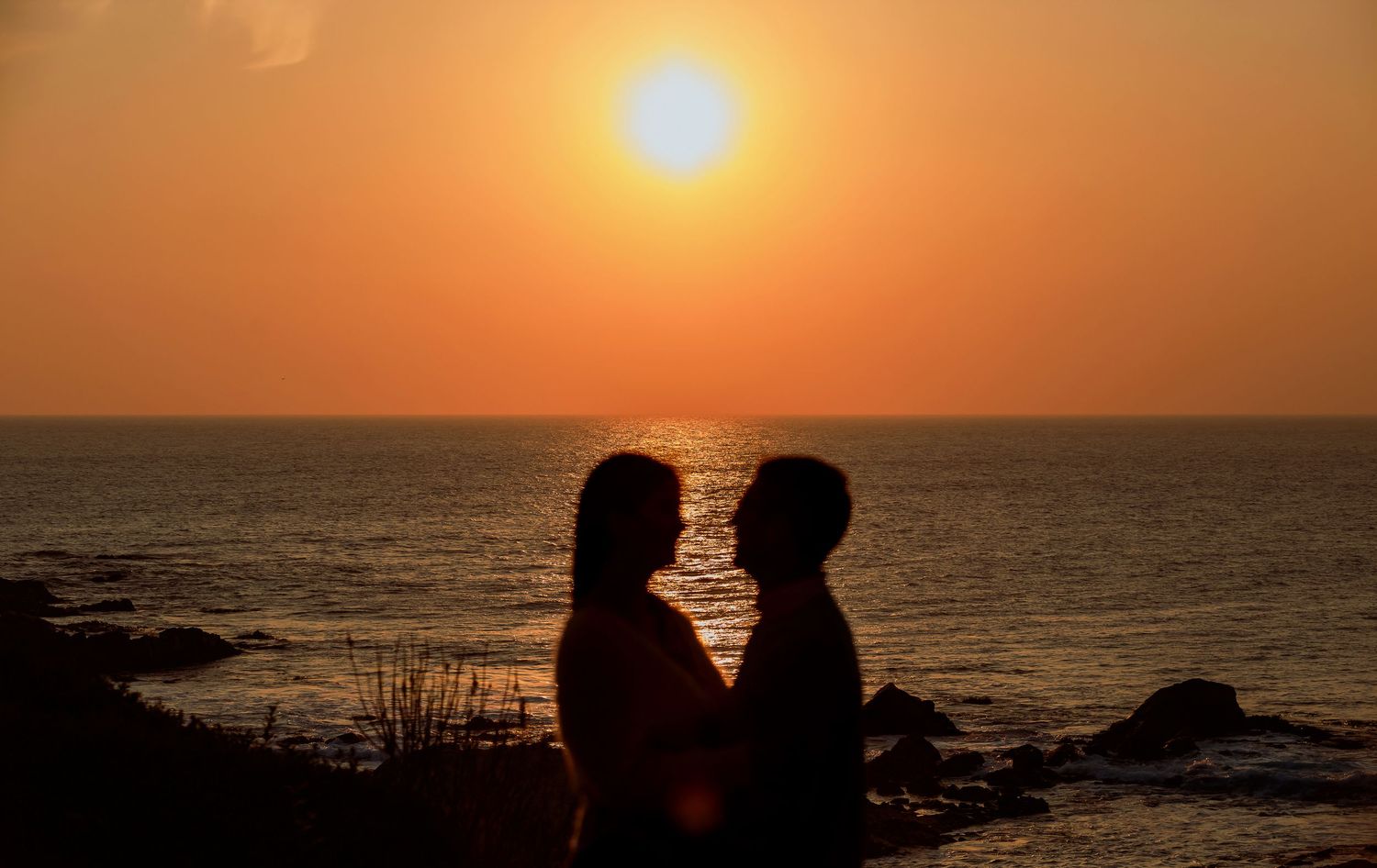 Silhouette of romantic couple facing each other against vibrant orange sunset over ocean horizon.