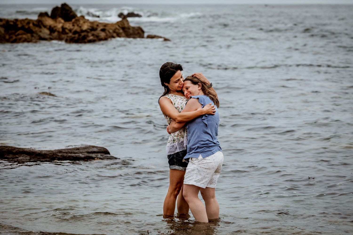 Two women embrace while standing in ocean waves at a rocky beach.