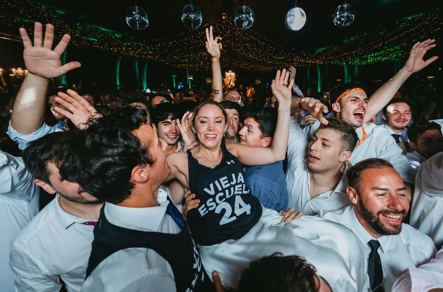 Bride in black tank top crowd surfing with guests at lively wedding reception.