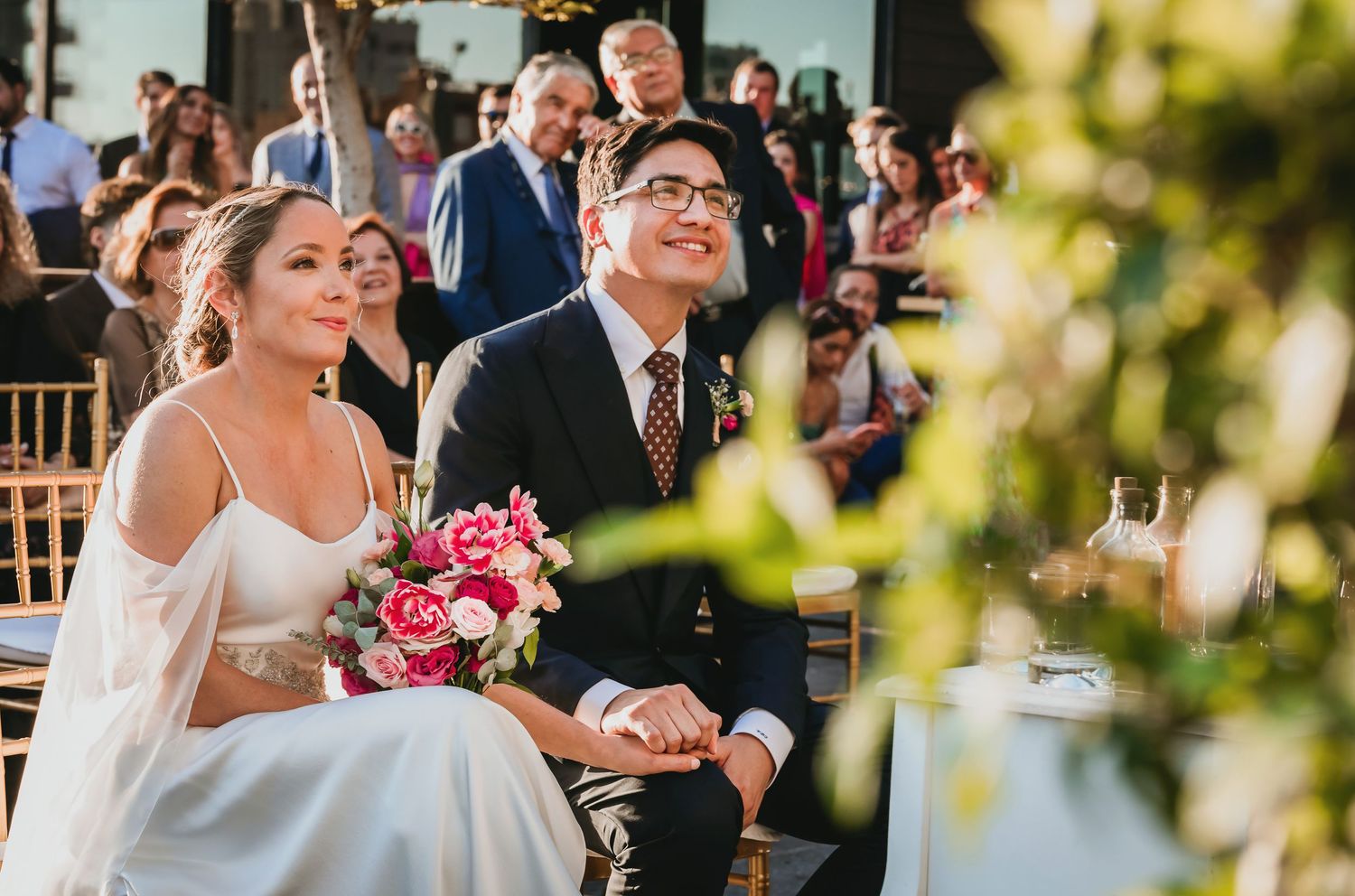 Bride and groom smile and hold hands during outdoor rooftop wedding ceremony with guests.