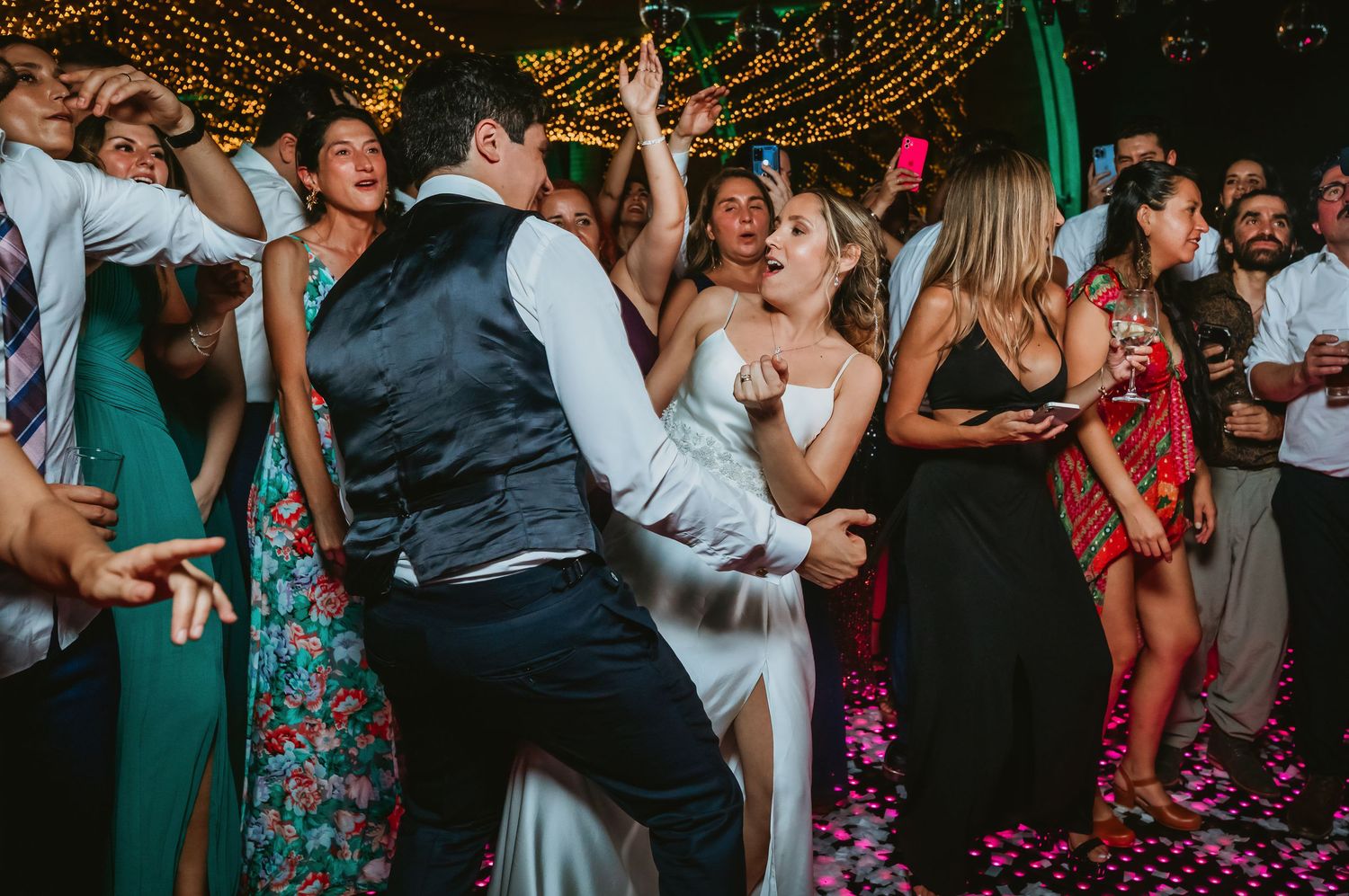 A bride in a white gown and groom dance energetically together surrounded by cheering guests at an outdoor wedding reception.