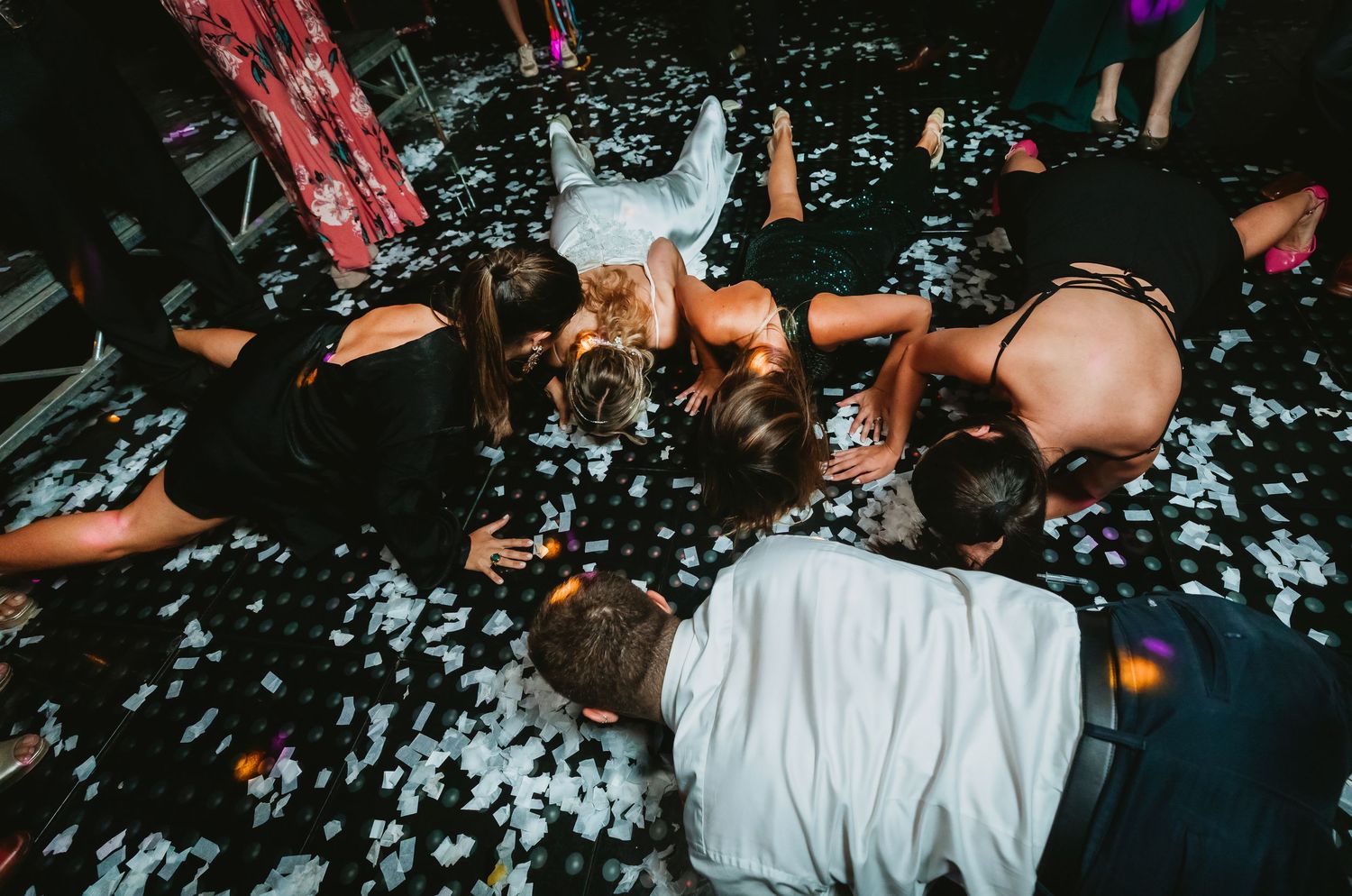 Wedding party lying on confetti-covered dance floor in fun celebration.