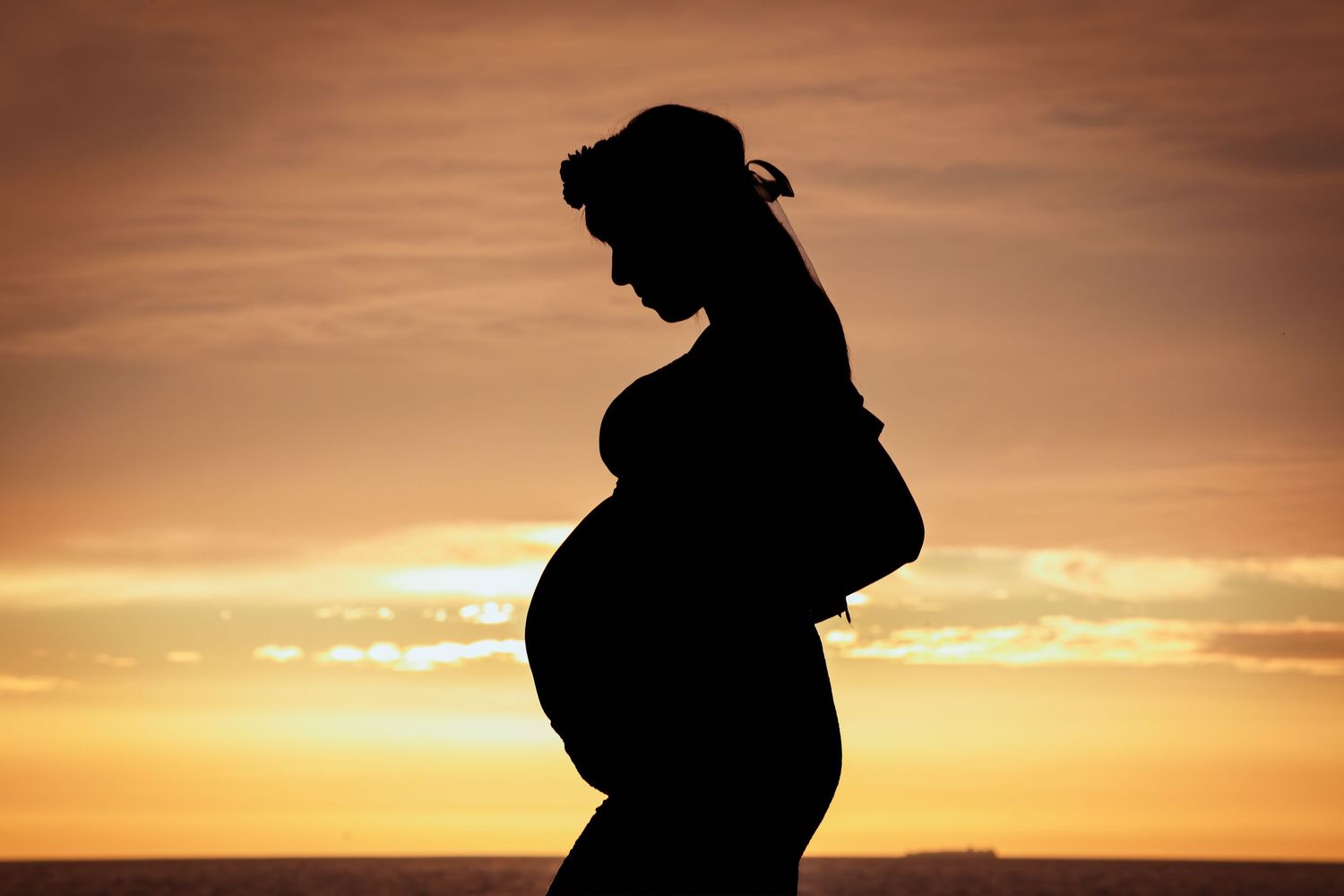 Silhouette of a pregnant woman cradling her baby bump against a warm golden sunset sky.