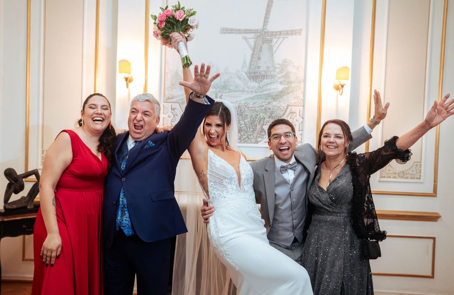 Joyful wedding celebration with bride in white gown, groom in gray suit, and three family members cheering indoors.