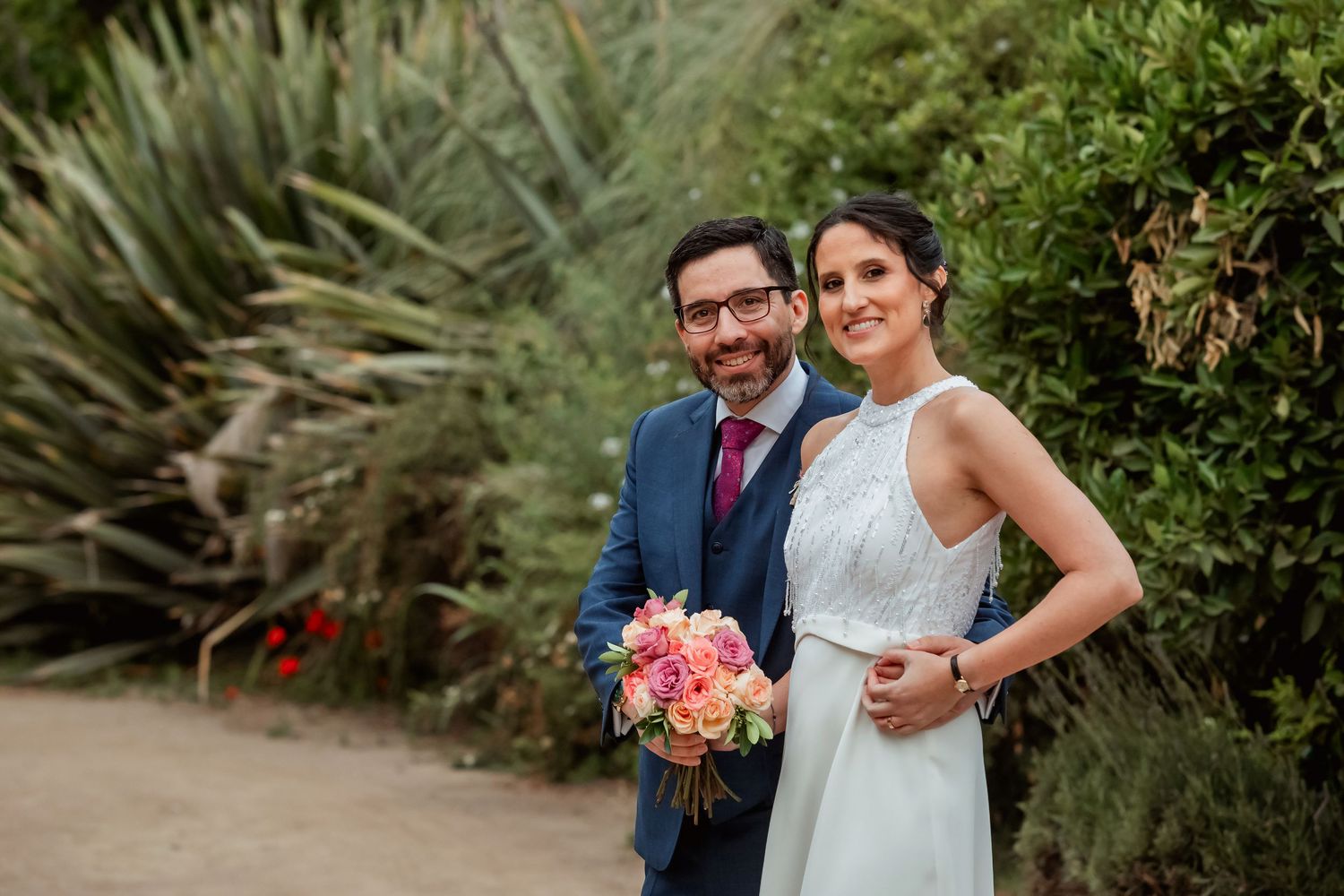 Bride and groom posing together in a garden, bride holding a colorful bouquet, groom in a navy blue suit.