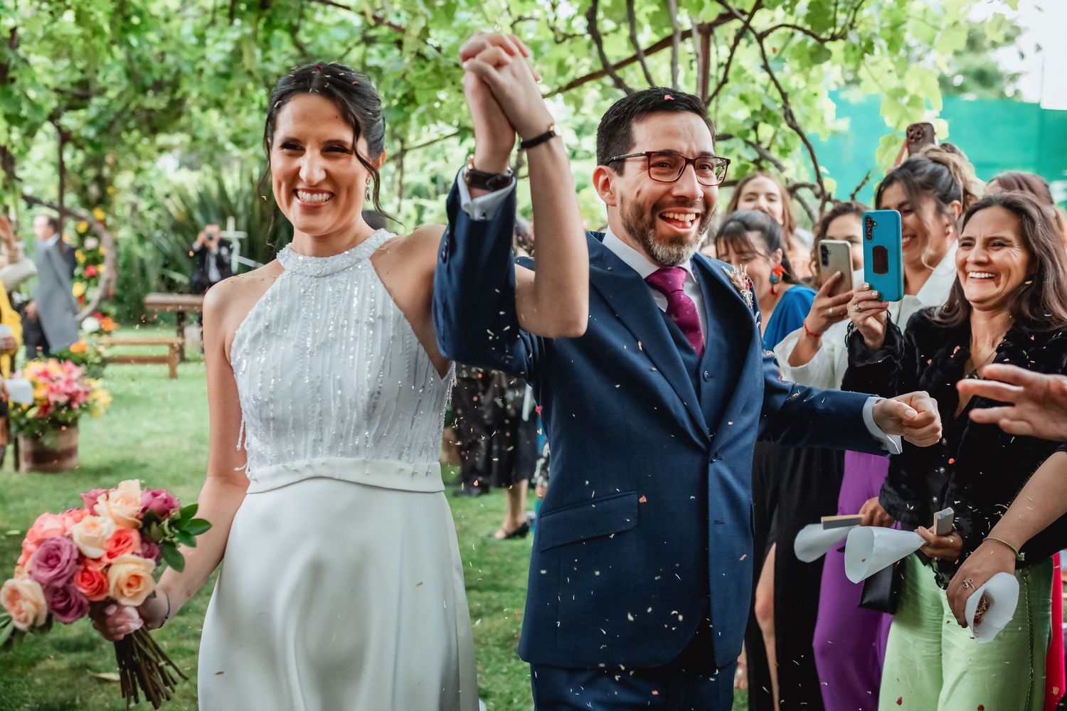 Newlyweds celebrate with raised hands walking through confetti after outdoor wedding ceremony.