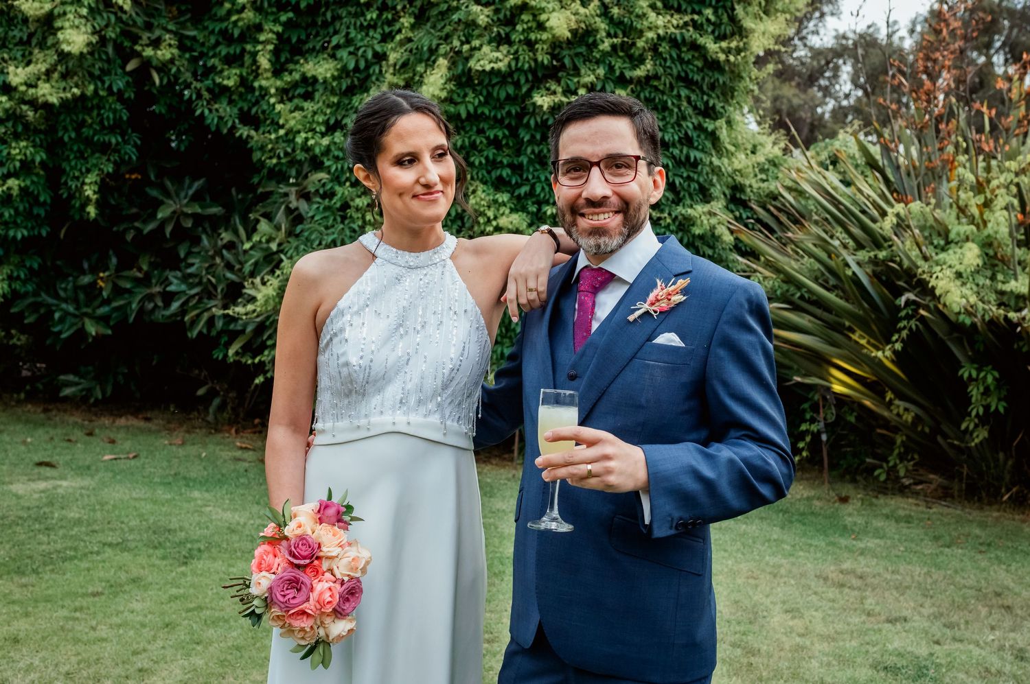 Smiling bride holding pink bouquet and groom with champagne pose in garden setting.