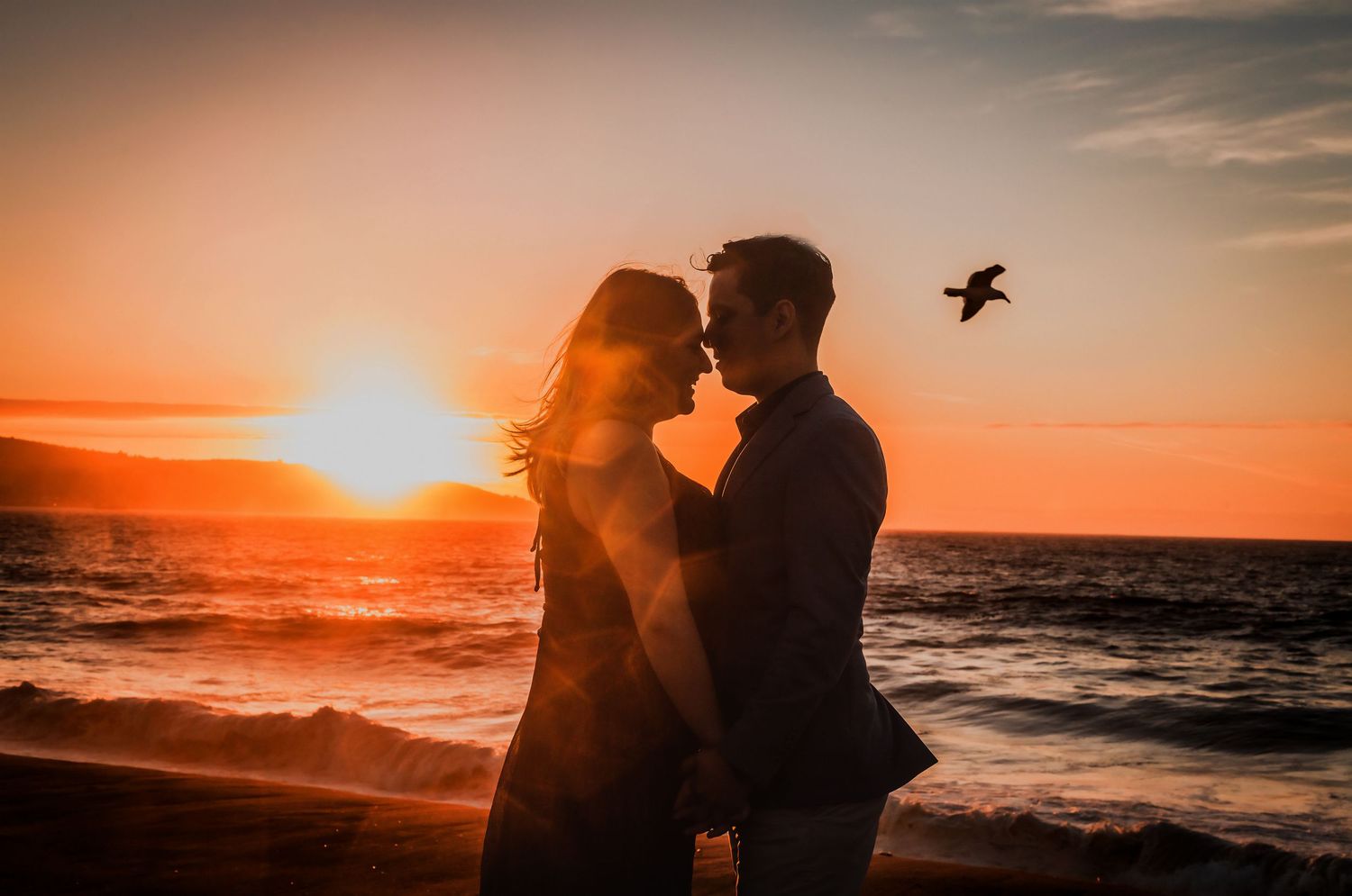 Romantic couple silhouetted against a vibrant ocean sunset with a bird flying overhead.
