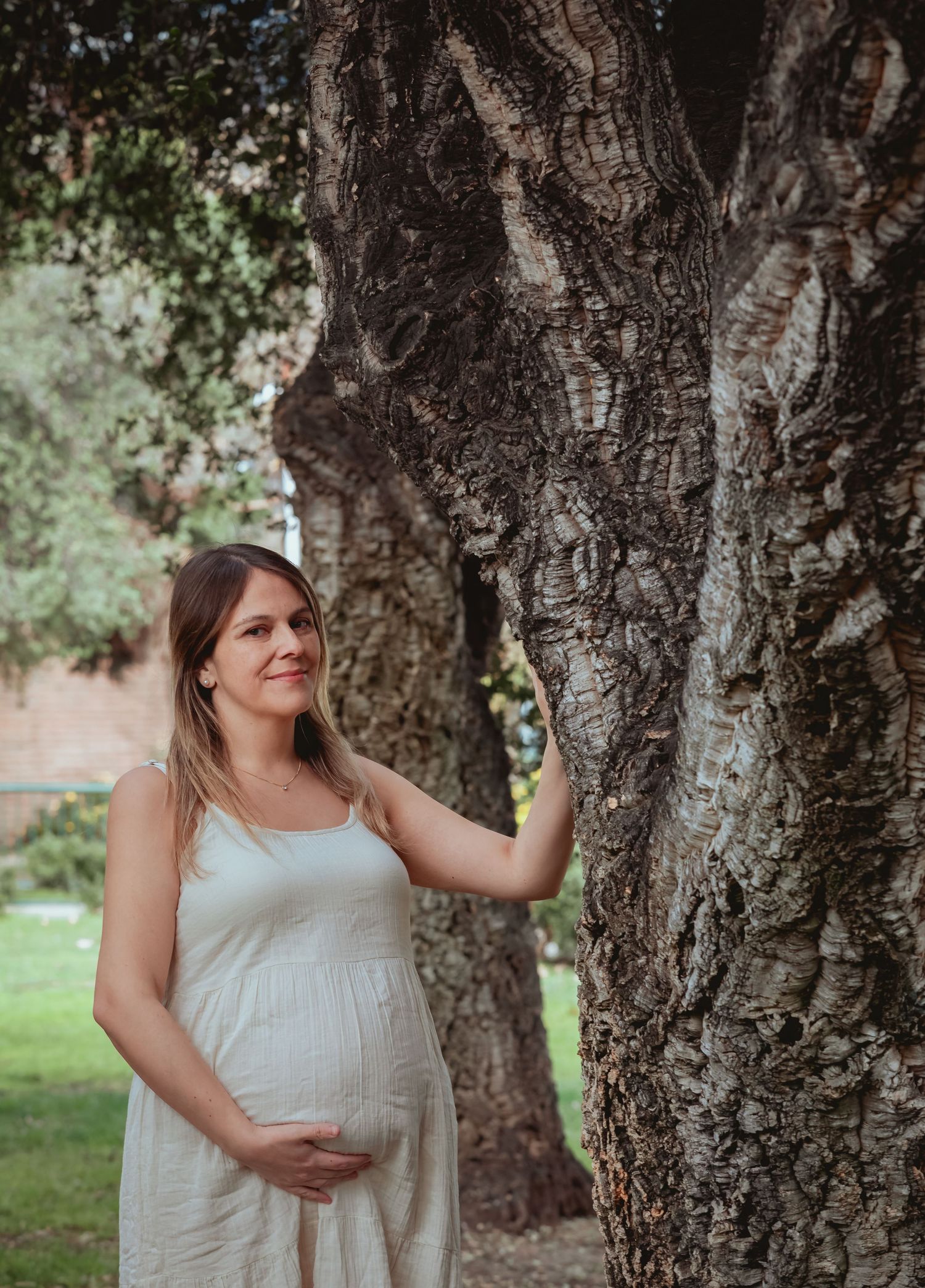 Pregnant woman in white dress leaning against a large textured tree trunk outdoors.