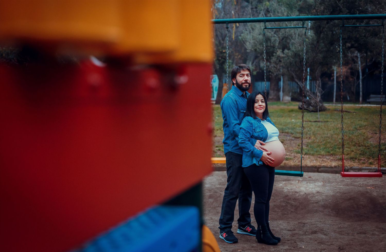 Pregnant couple embracing at a colorful playground in a park, man hugging woman from behind.