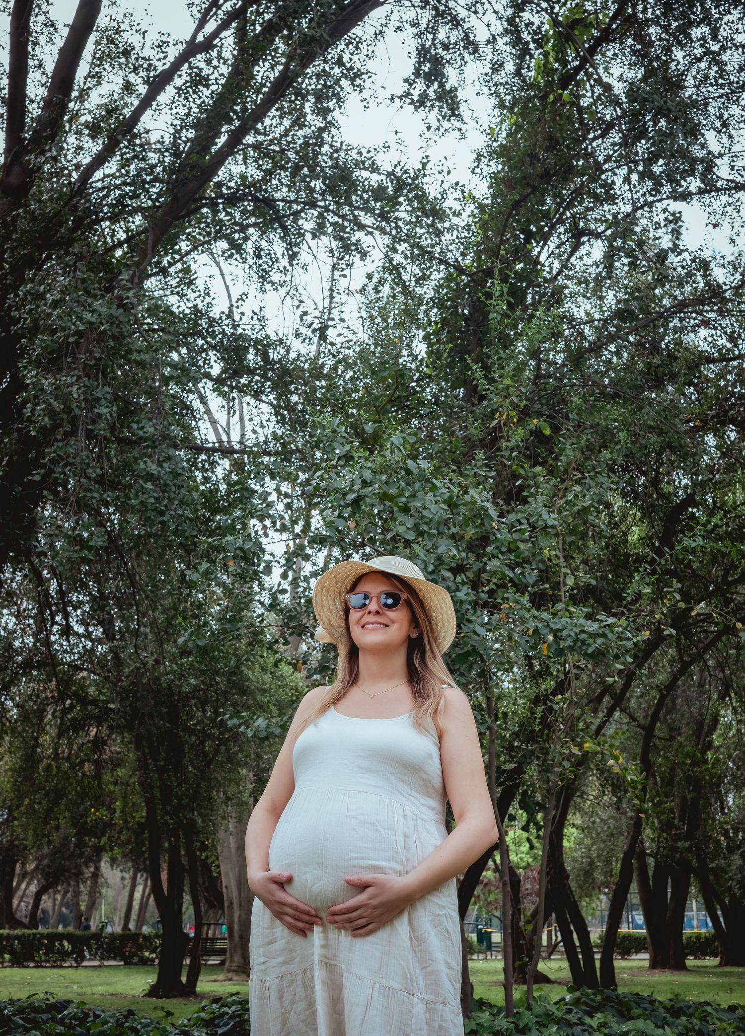 Pregnant woman in white dress and straw hat cradling belly among tall park trees.