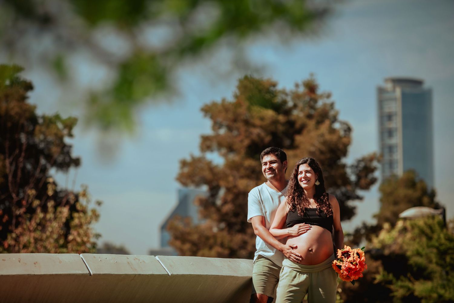 Couple smiling together outdoors with colorful flowers, lush trees, and a city skyscraper in background.