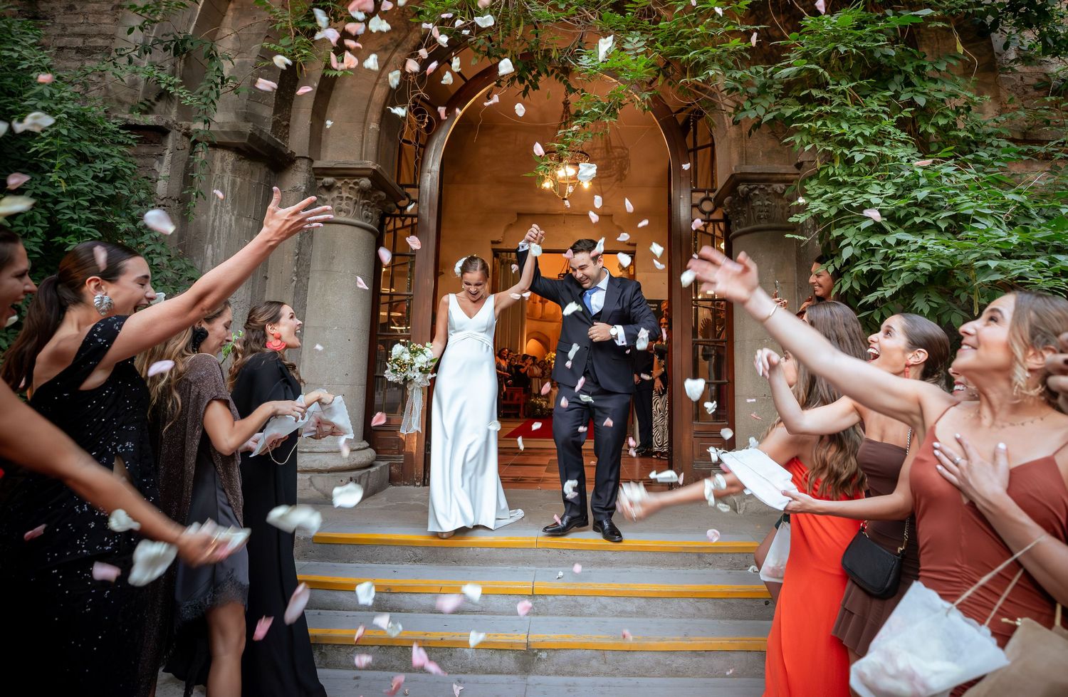 Bride and groom celebrate outside ornate church doorway as guests toss white flower petals joyfully.