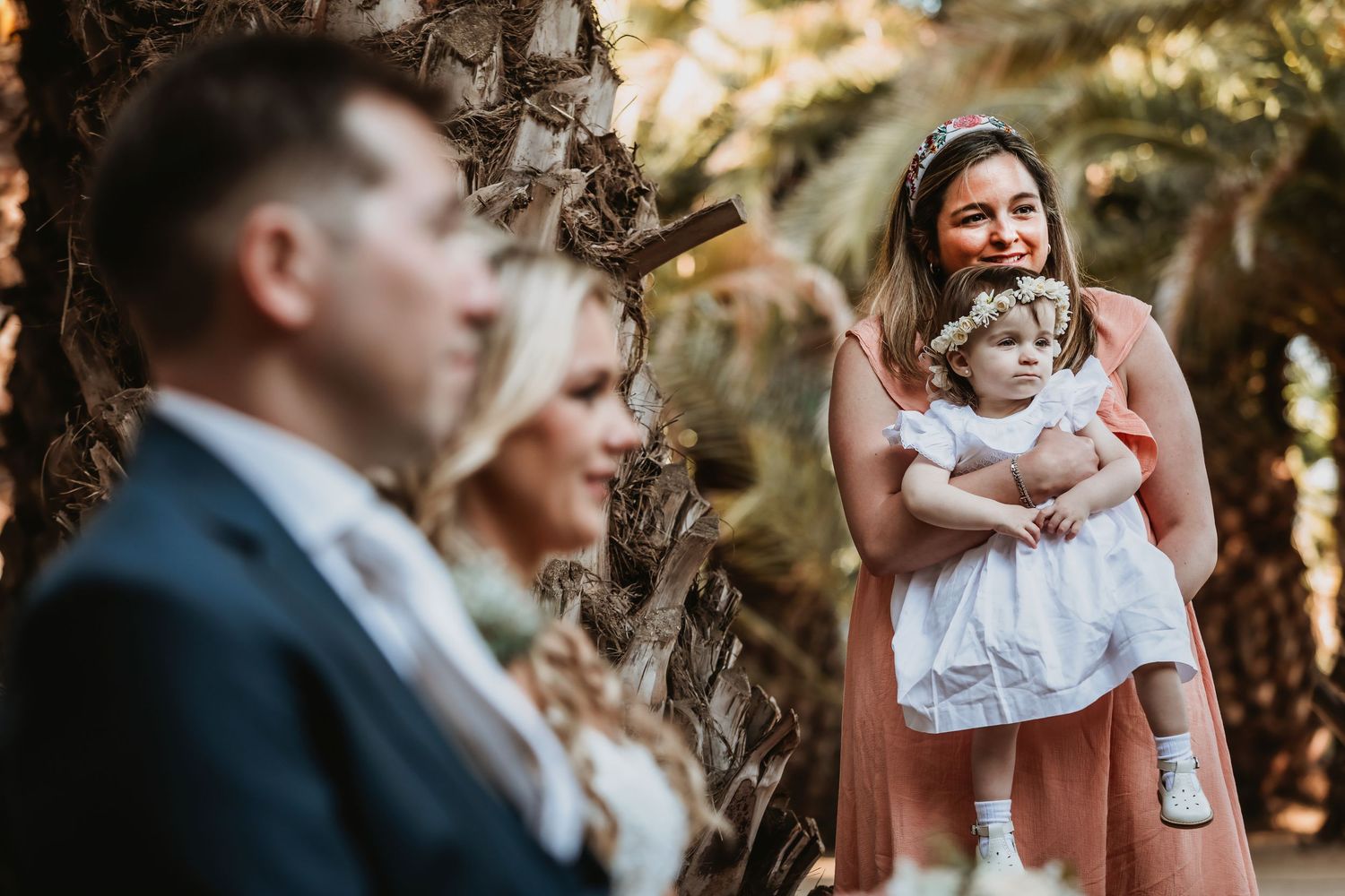 A woman in a pink dress holds a baby girl in white at an outdoor wedding ceremony among palm trees.