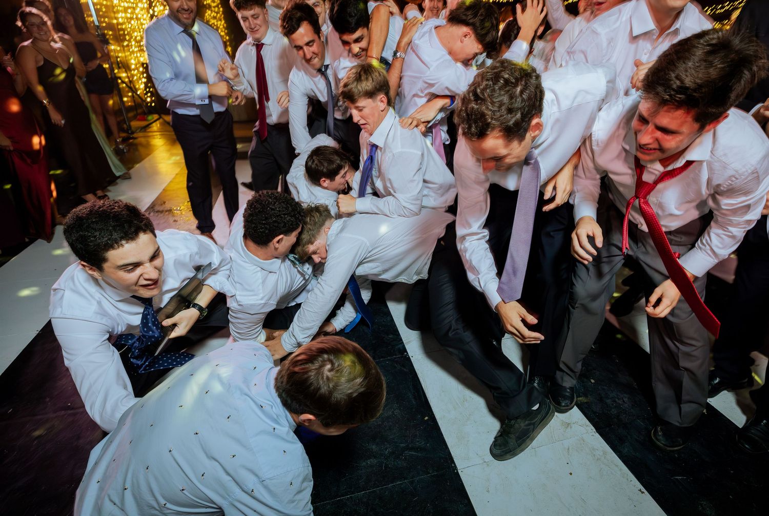 Young men in dress shirts pile onto dance floor in playful heap at wedding reception.