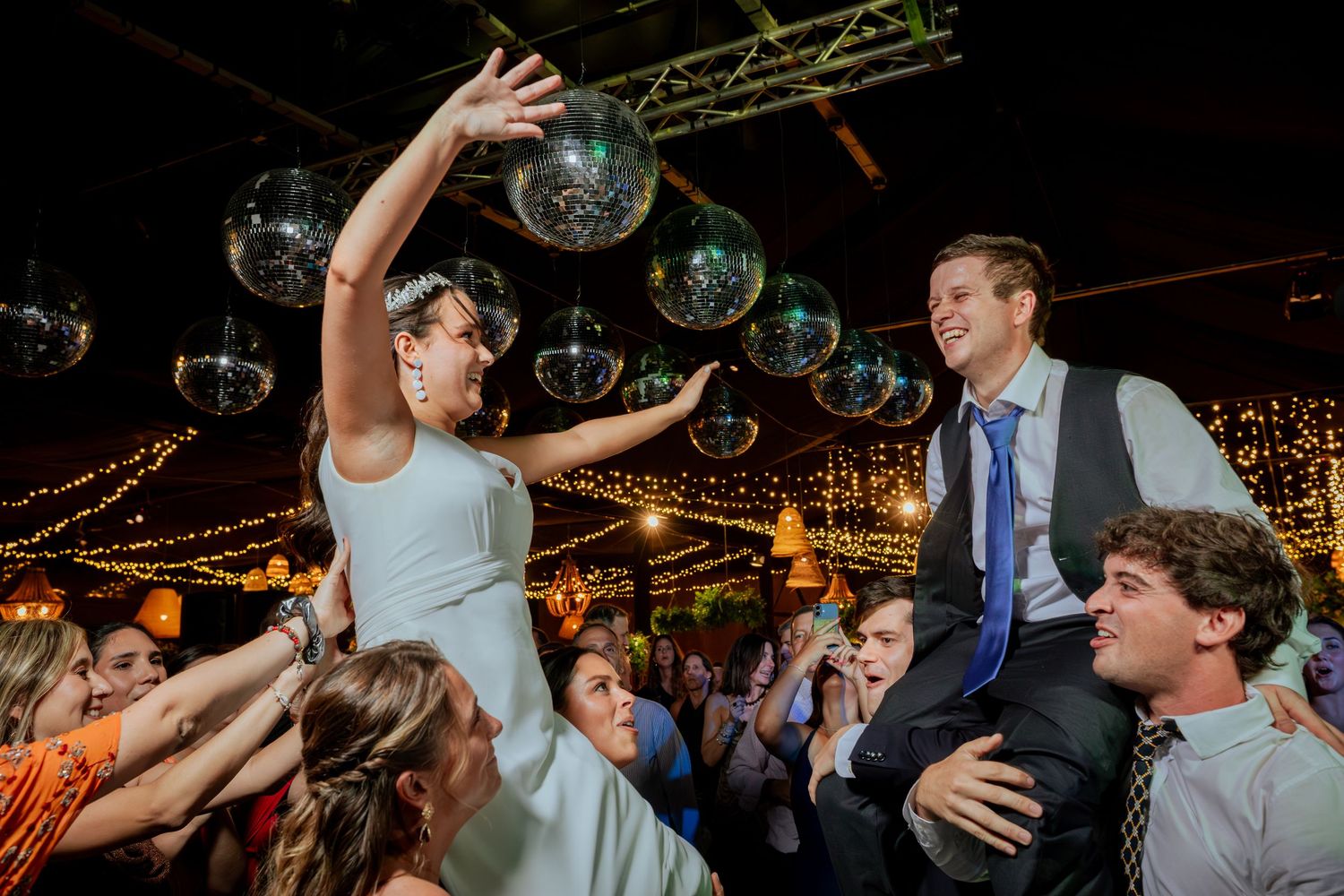 Bride lifted on shoulders reaching for disco ball at lively indoor wedding reception party.