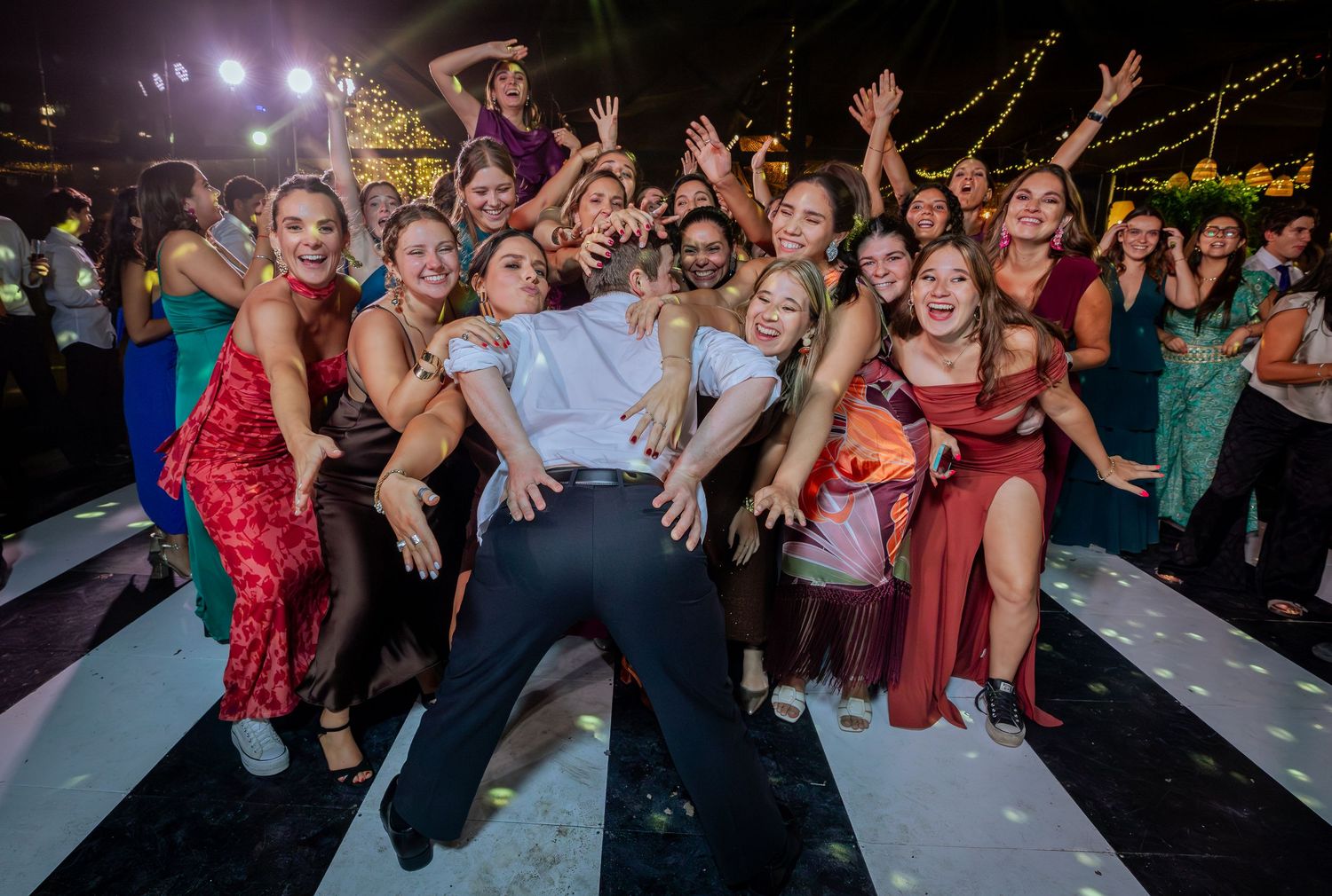 Groom surrounded by cheering female wedding guests on dance floor at reception.