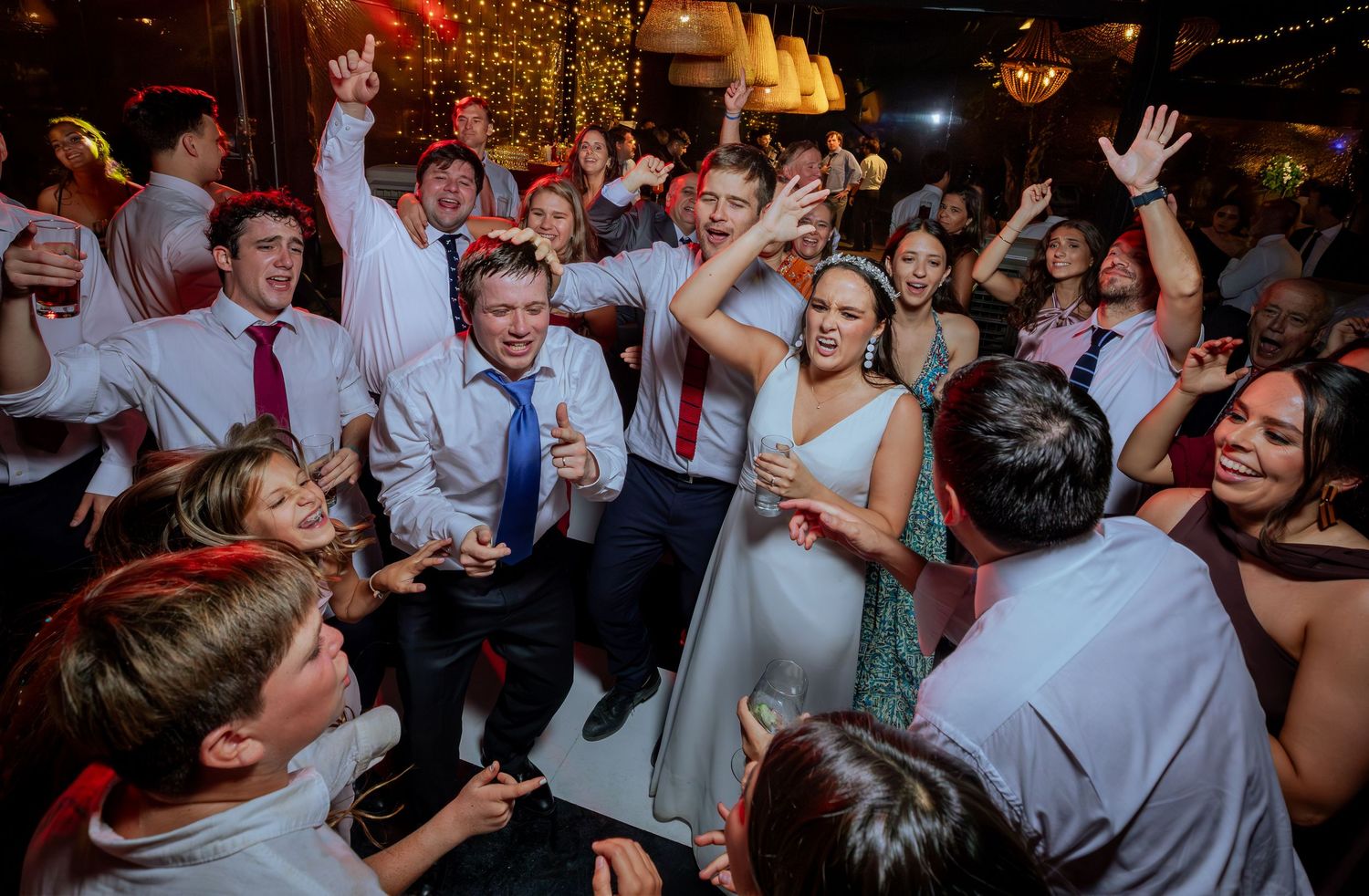 Bride in white gown celebrates with energetic dancing guests at festive wedding reception party.