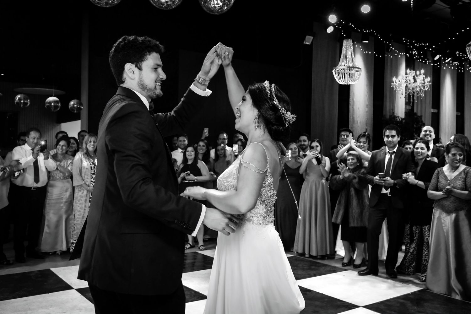 Newlyweds dance together on checkered floor as smiling wedding guests clap and celebrate around them.