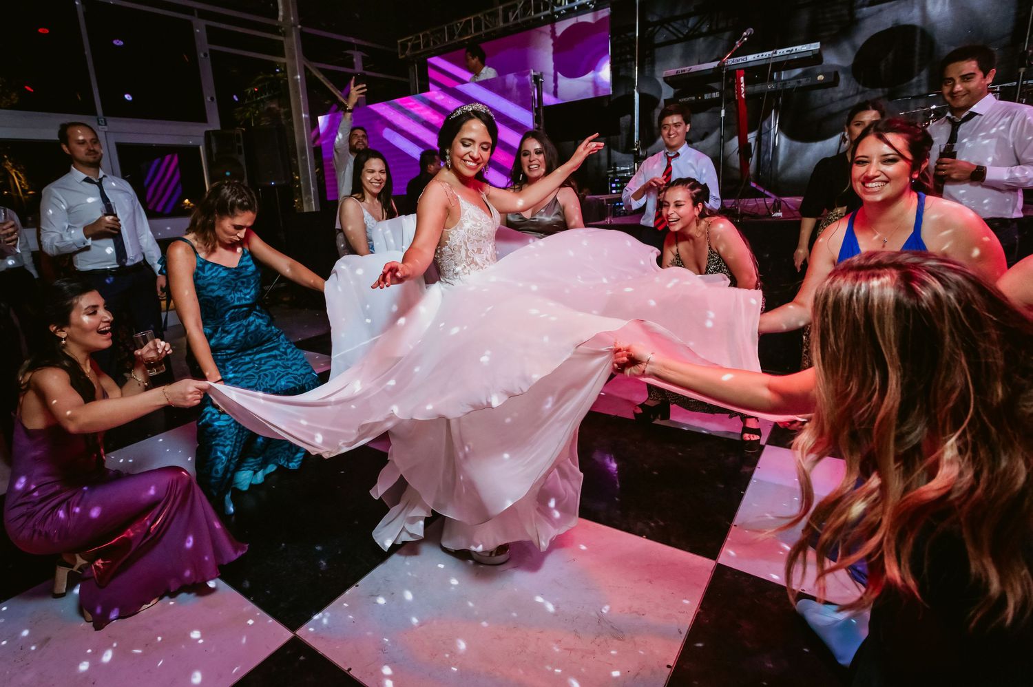 Bride in elegant white gown spins on checkered dance floor as guests cheer at lively wedding reception.