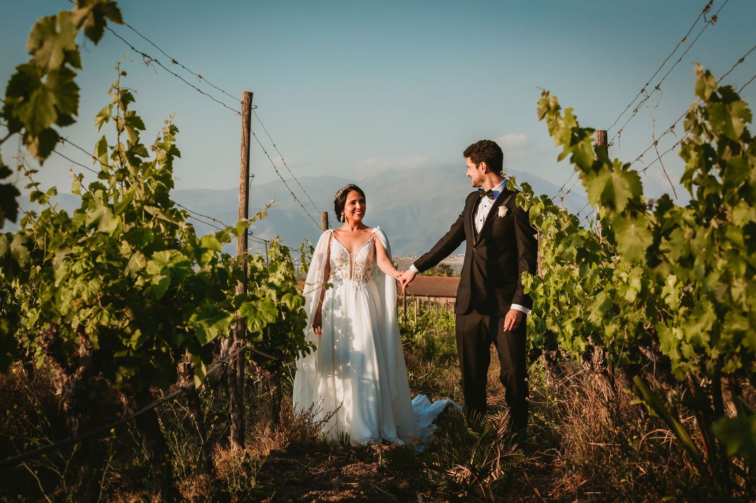 Newlyweds hold hands and walk through a vibrant green vineyard under a clear blue sky.