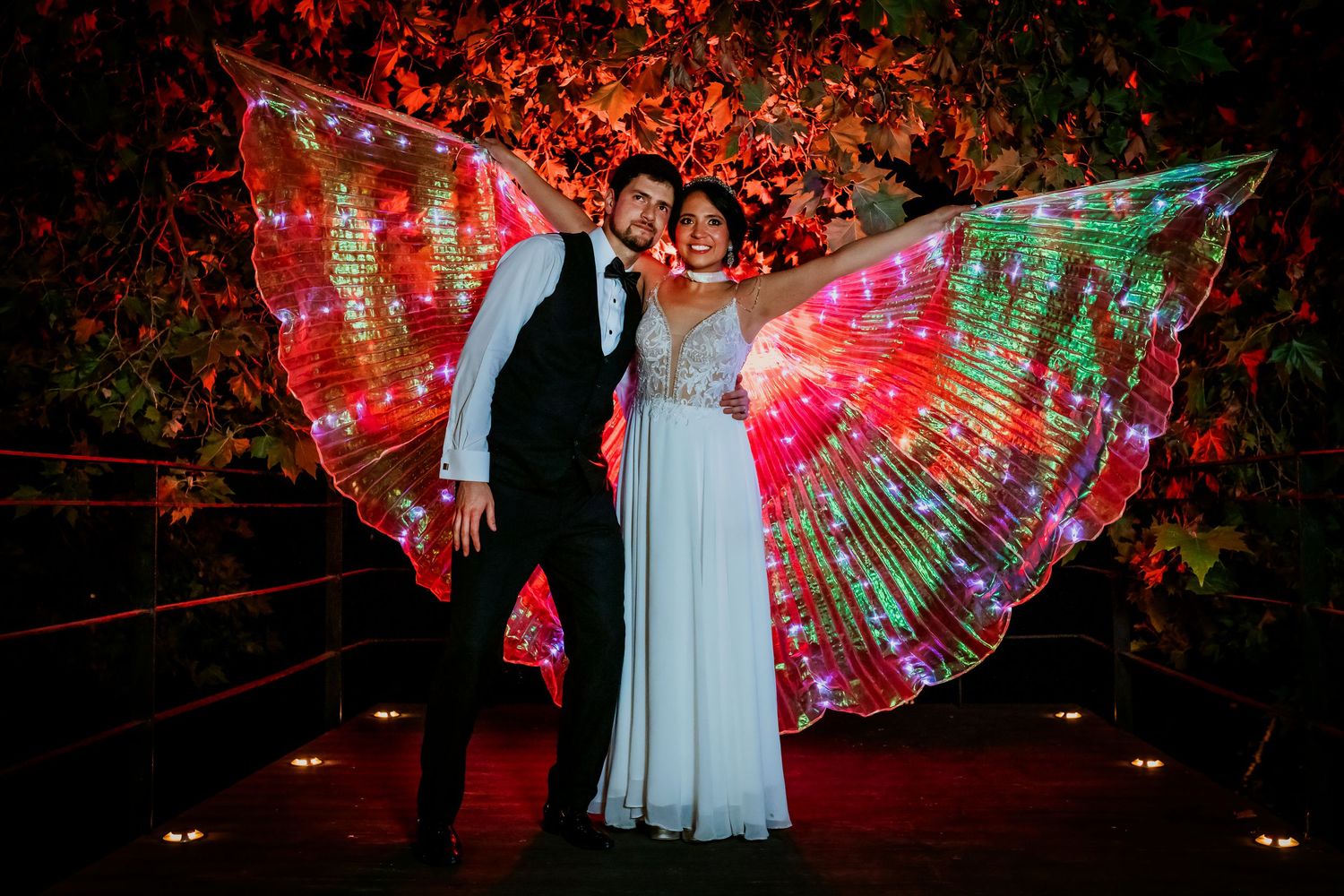 Couple posing outdoors at night with bride wearing vibrant illuminated red and green LED wings.