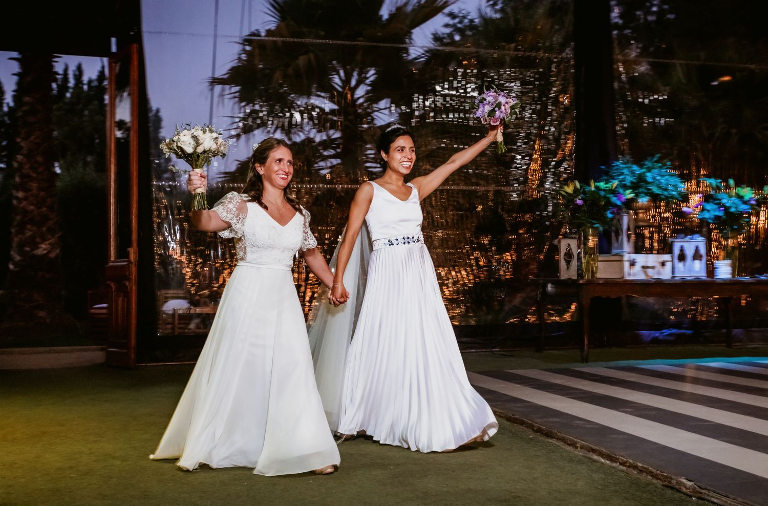 Two brides hold hands and raise bouquets triumphantly at their outdoor evening wedding reception.