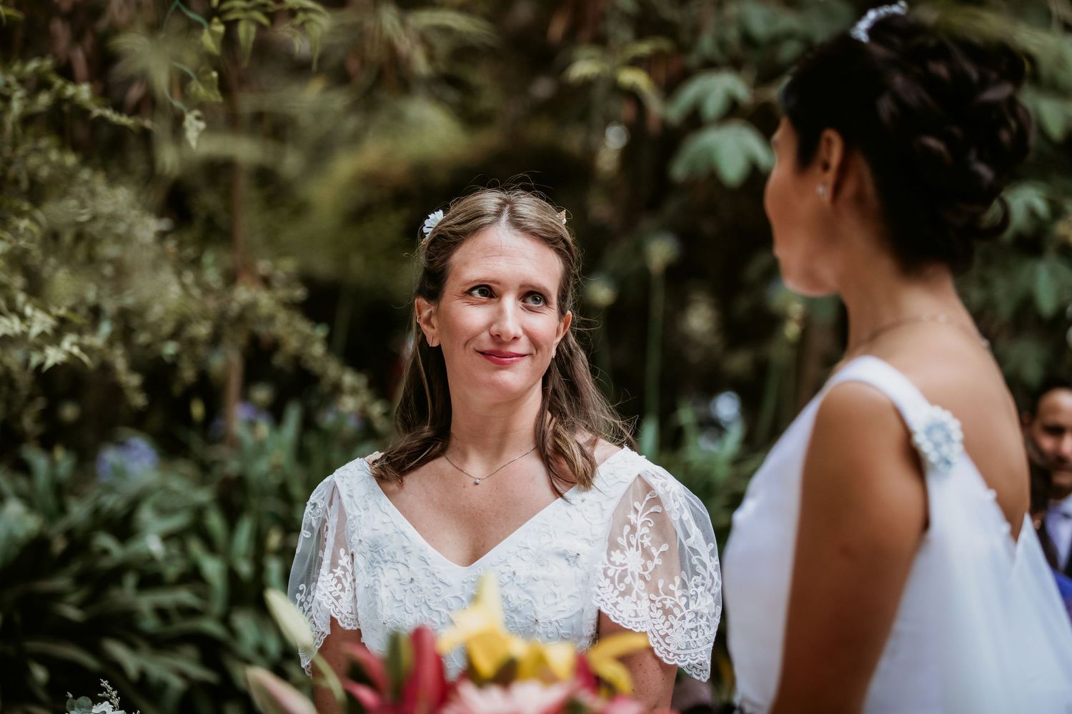 Bride in lace dress gazes lovingly at her partner during outdoor wedding ceremony.