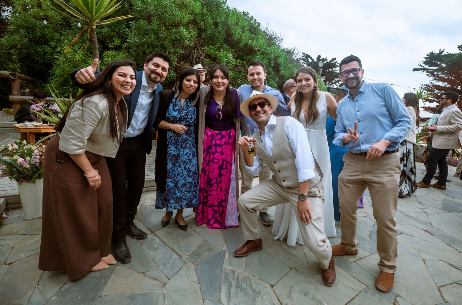 Group of smiling wedding guests posing together on a stone patio at an outdoor celebration.