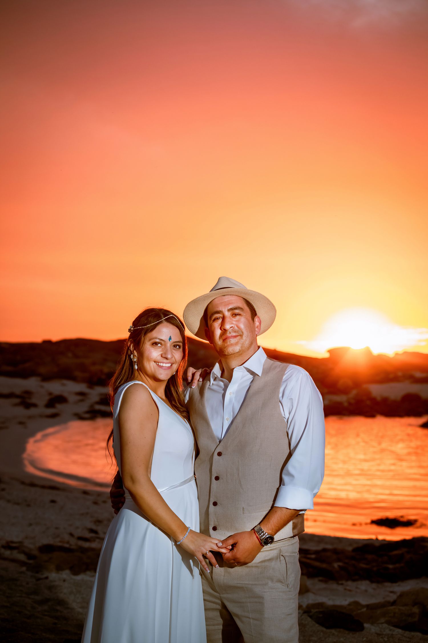 Bride and groom holding hands at sunset on a rocky beach, smiling warmly in wedding attire.