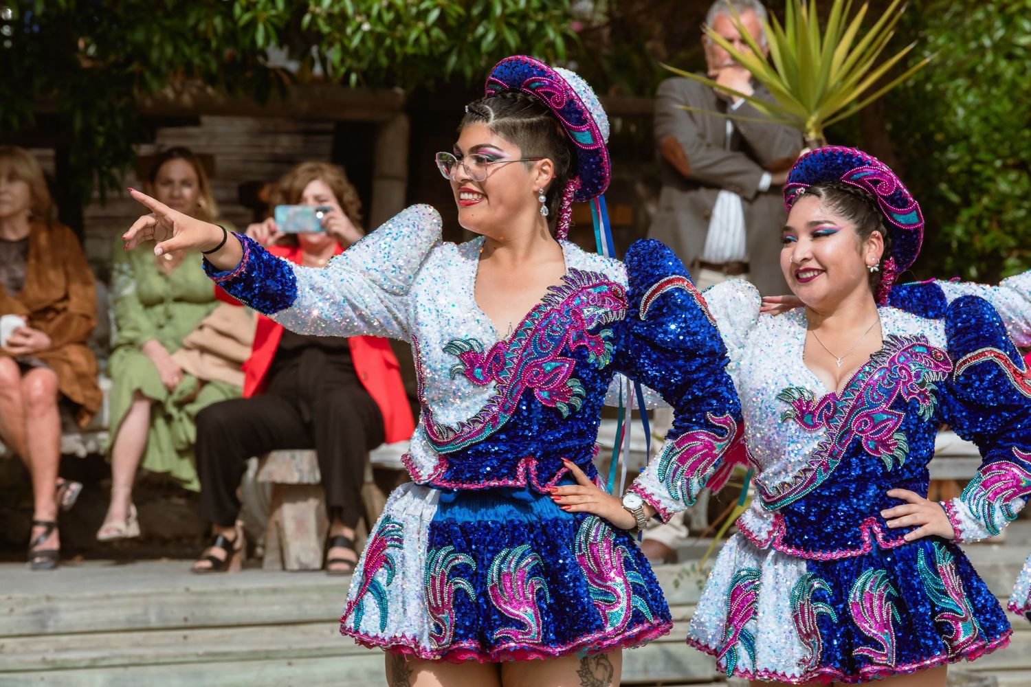 Two women in vibrant blue and pink traditional embroidered costumes perform folk dance at outdoor wedding.