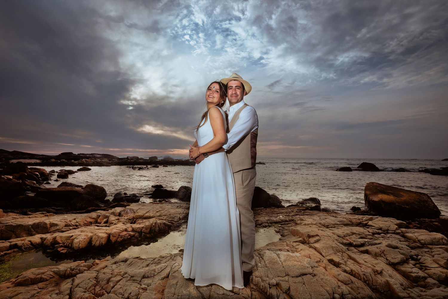 Bride and groom embracing on scenic rocky coastline with dramatic sunset clouds above.