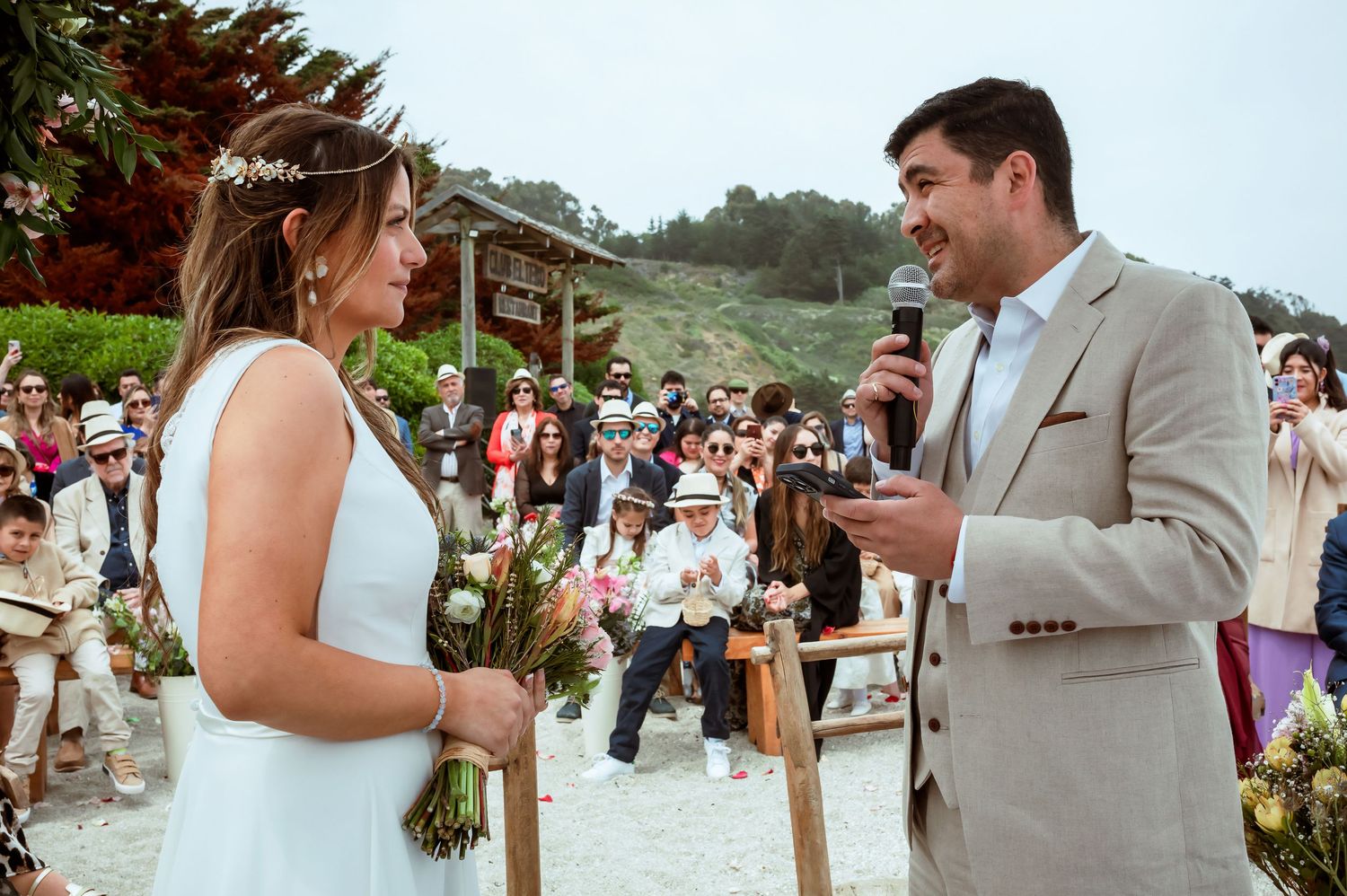 Groom speaks into microphone facing bride during outdoor beach wedding ceremony with guests behind.