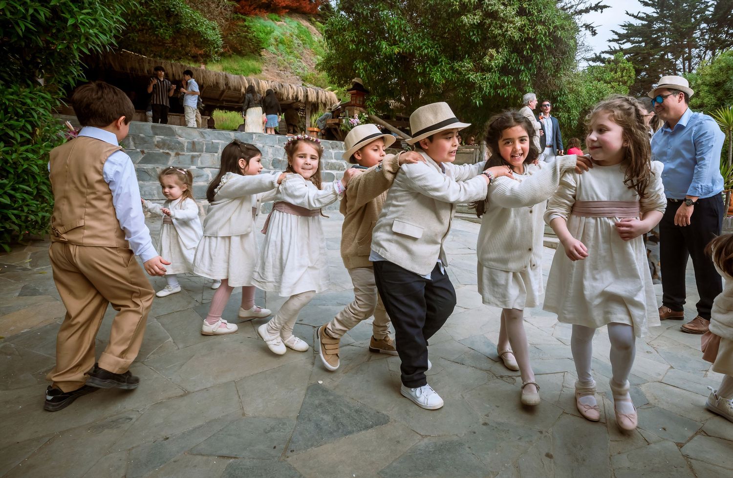 Adorable children in formal wedding attire forming a dancing chain on a stone patio at an outdoor wedding.