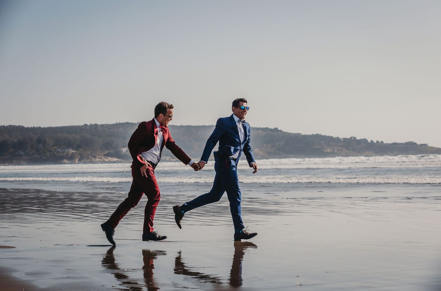 Two grooms in red and blue suits running hand in hand along a reflective beach shoreline.
