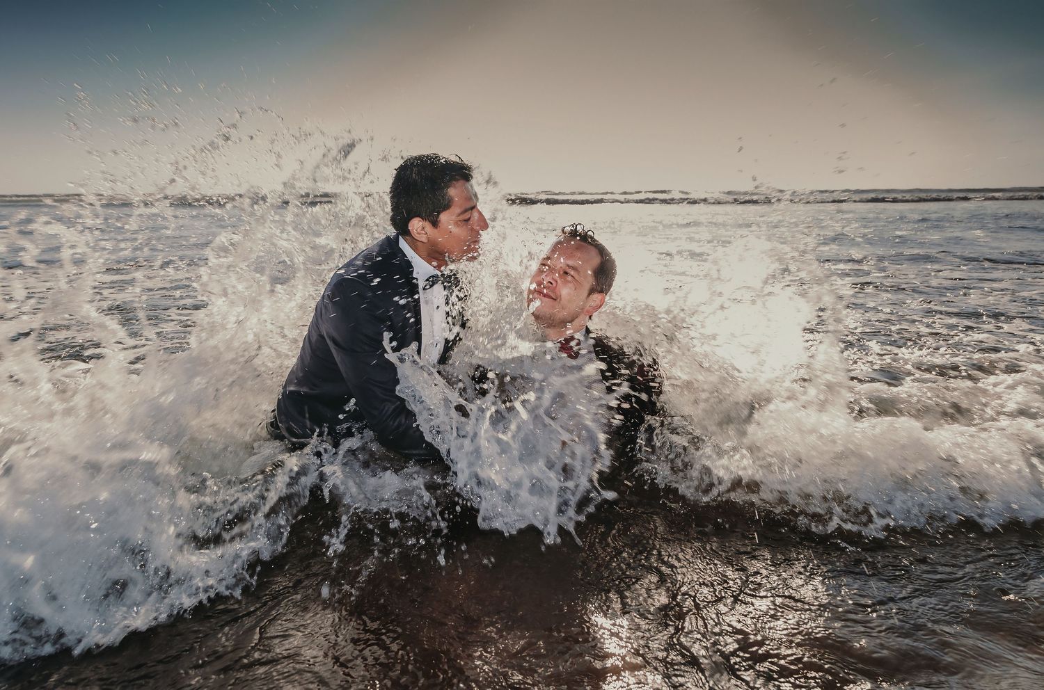 Two grooms in tuxedos embrace as a large ocean wave crashes over them on their wedding day.